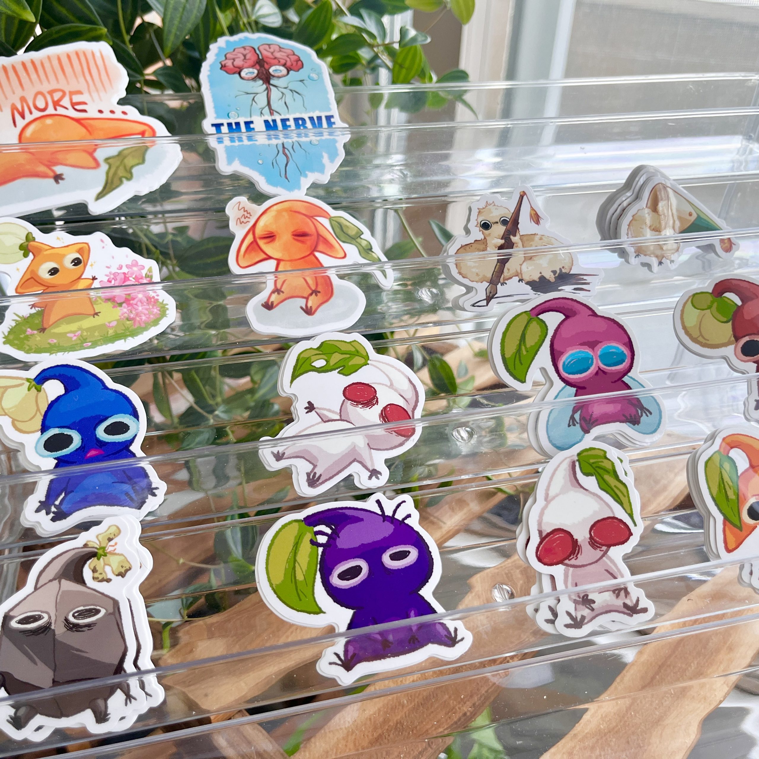 Display of colourful Pikmin-inspired stickers arranged in clear trays, with small plant-like characters, fruit-themed creatures, and a “The Nerve” sticker.