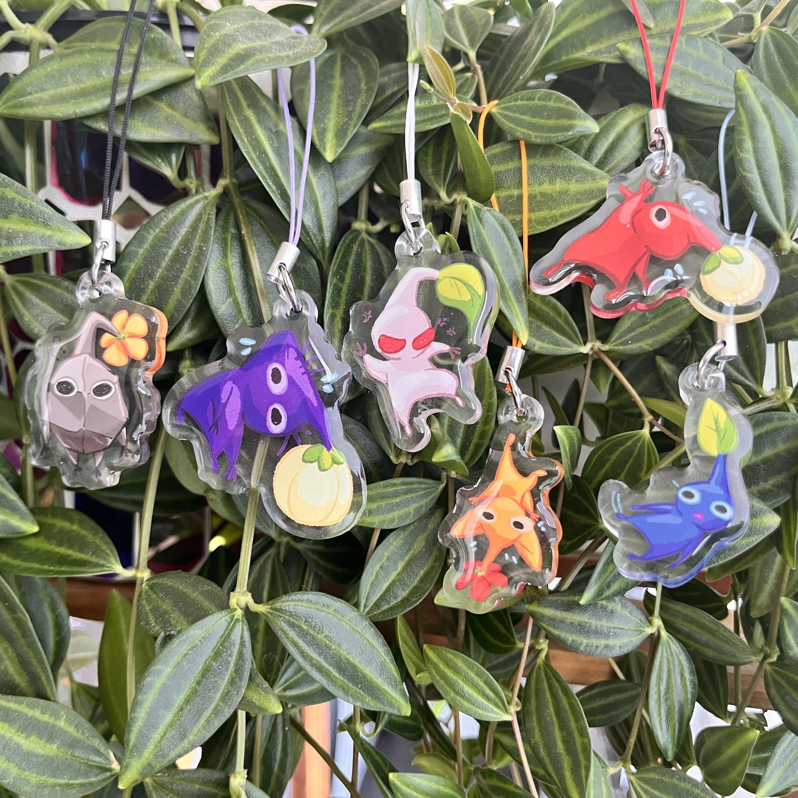 Acrylic phone charms of Pikmin-inspired characters hanging among green houseplant leaves.