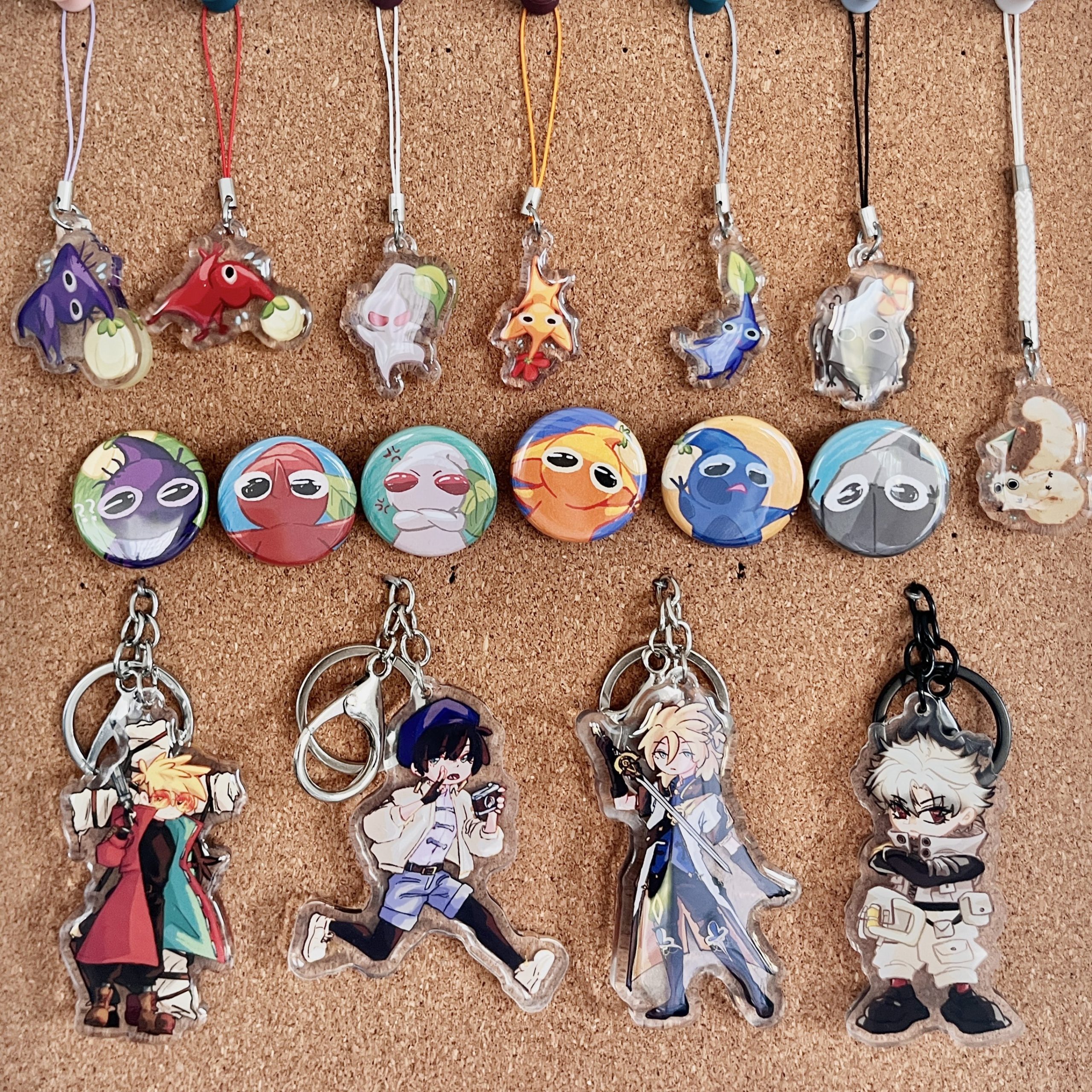 Corkboard display of acrylic charms, buttons, and keychains featuring Pikmin-inspired characters along with anime and video game character charms.