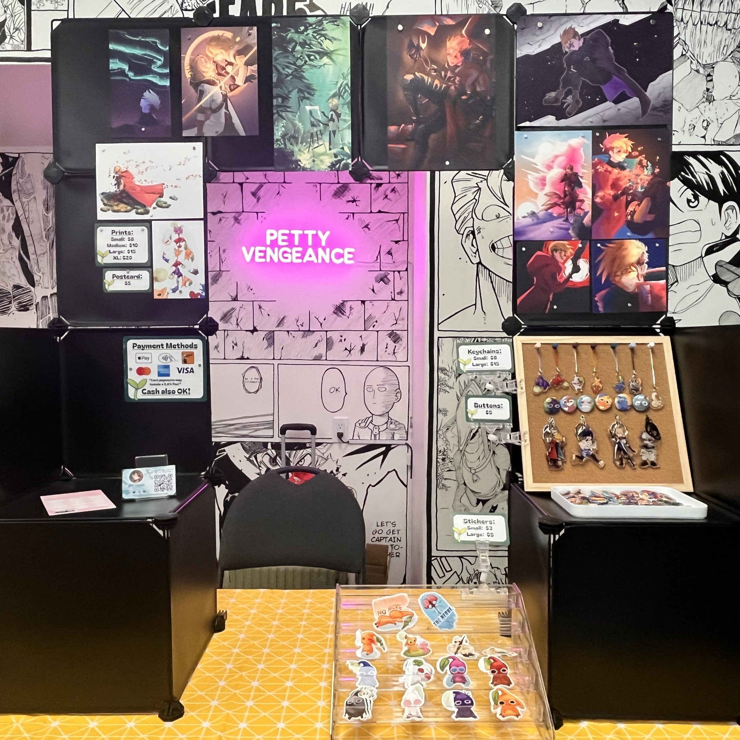 Artist alley table display with anime and video game prints, stickers, buttons, charms, and keychains, including a pink “Petty Vengeance” sign at the centre.