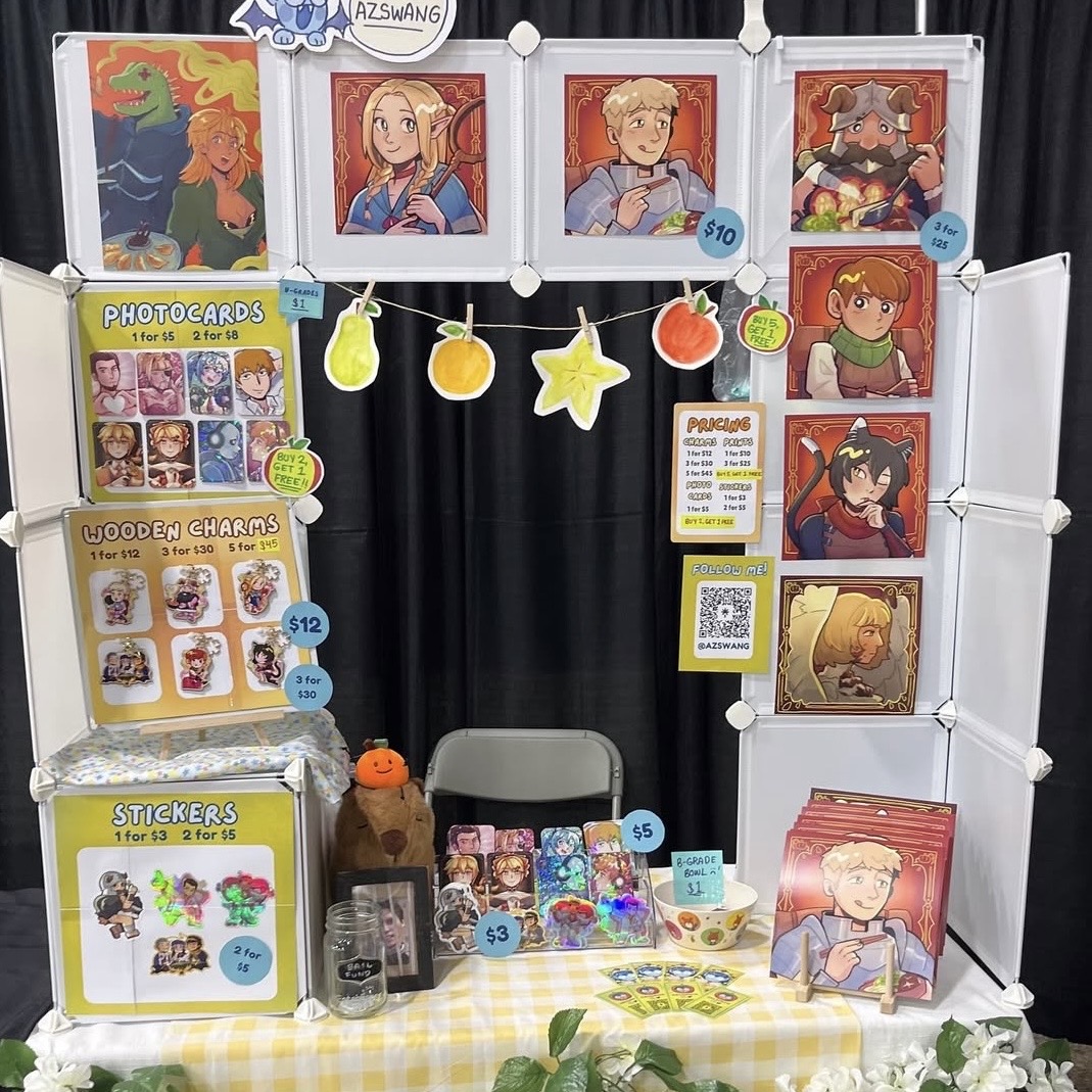 Artist alley booth display with colourful anime, game, and comic-inspired prints, photocard sets, wooden charms, stickers, and price signs.