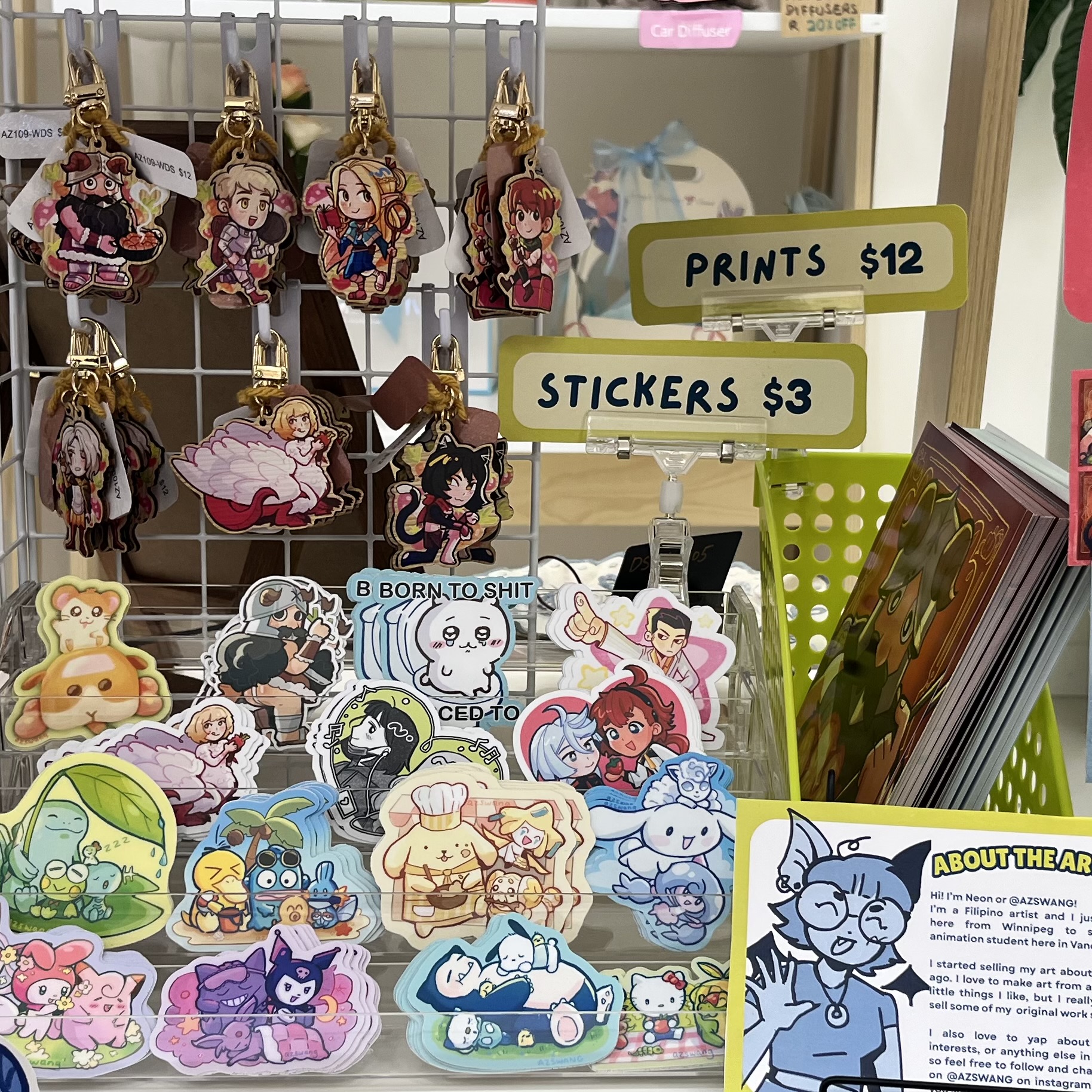 Close-up of AZSWANG merchandise including wooden charms, colourful stickers, art prints, price signs, and an “About the Artist” card.