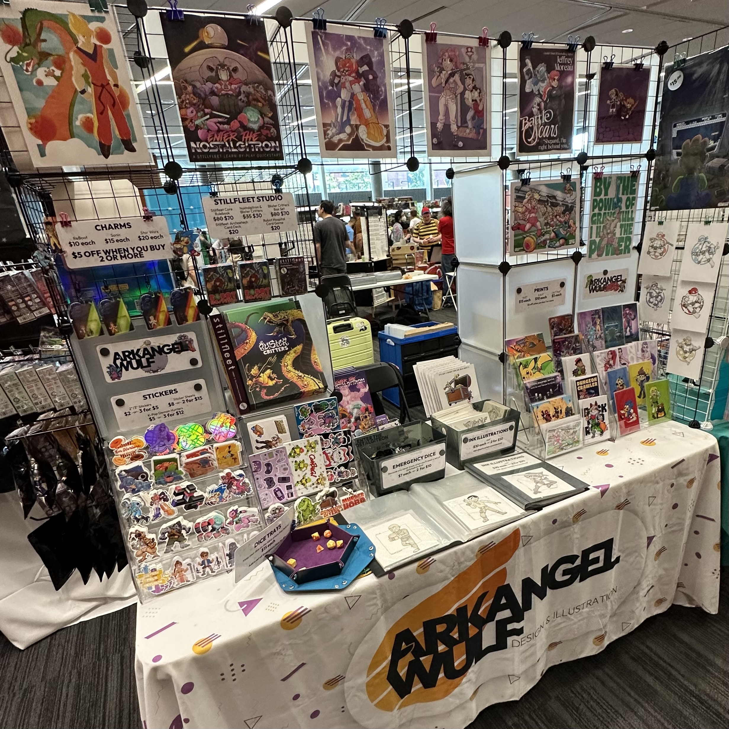 Arkangel Wulf vendor table displaying fandom-inspired prints, stickers, charms, roleplaying game books, dice trays, art portfolios, and Stillfleet Studio game materials.
