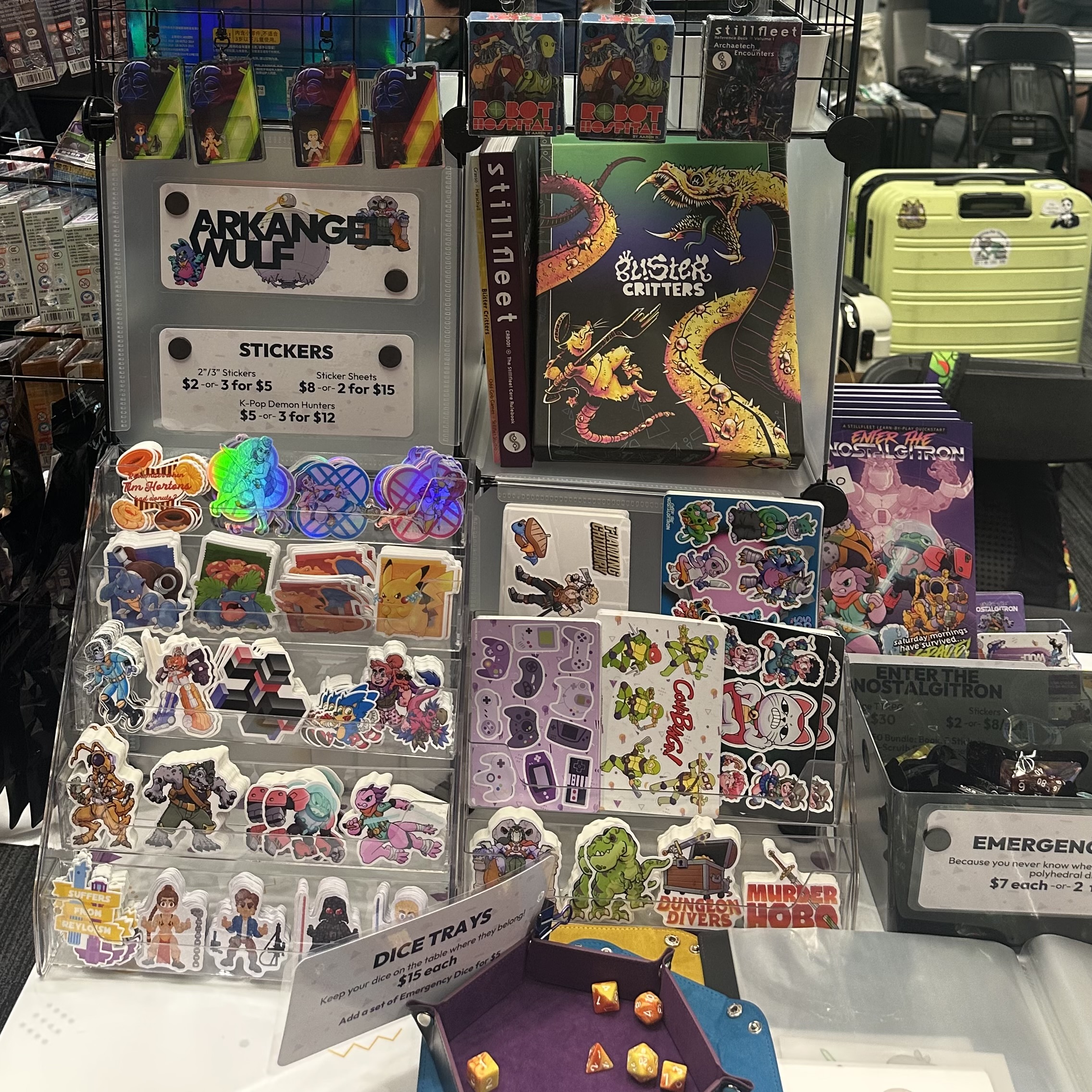 Close-up of Arkangel Wulf merchandise including colourful fandom stickers, sticker sheets, charms, dice trays, emergency dice, and Stillfleet Studio books and game materials.