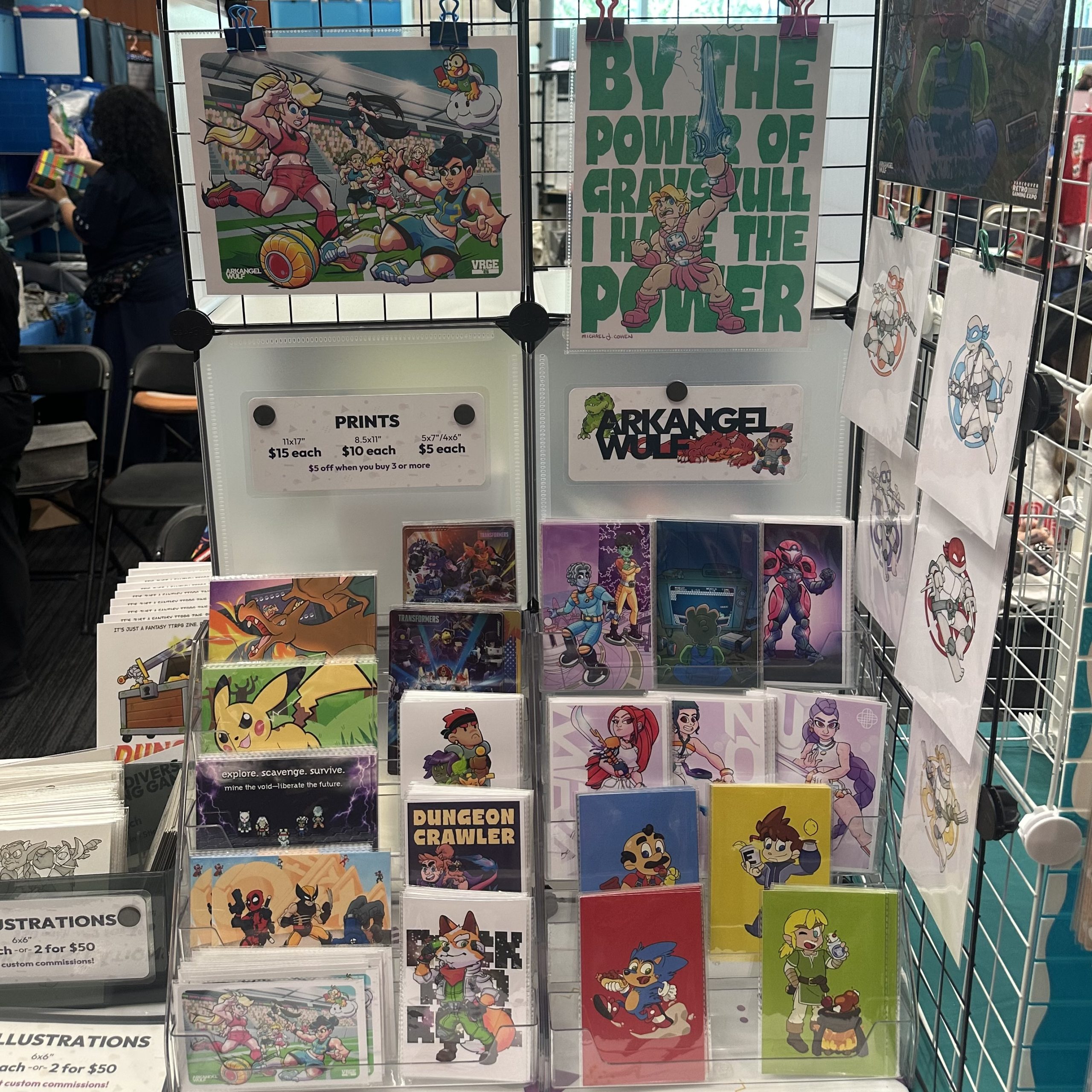 Display of Arkangel Wulf art prints and postcards featuring video game, fantasy, and pop culture-inspired characters, with price signs and illustrated robot designs hanging beside them.