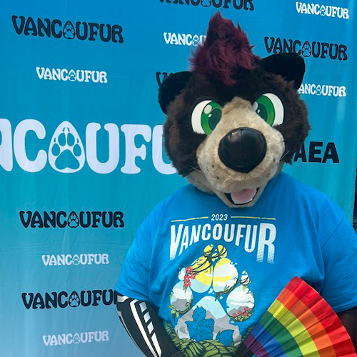 Fursuiter wearing a blue Vancoufur 2023 T-shirt and holding a rainbow fan in front of a Vancoufur photo backdrop.