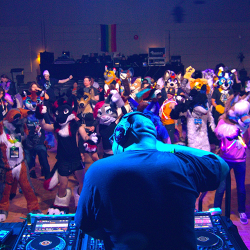 Crowd of fursuiters and attendees dancing at a furry convention event while a DJ plays music from the foreground.