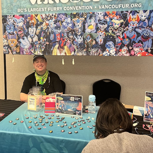 Vancoufur information table with buttons, prize items, and a volunteer seated beneath a banner showing a large group of fursuiters.