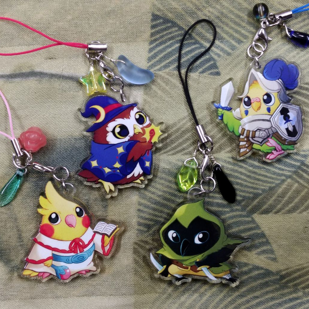 Four acrylic bird-themed charms on patterned fabric, featuring cute fantasy birds dressed as a mage, cleric, rogue, and warrior.