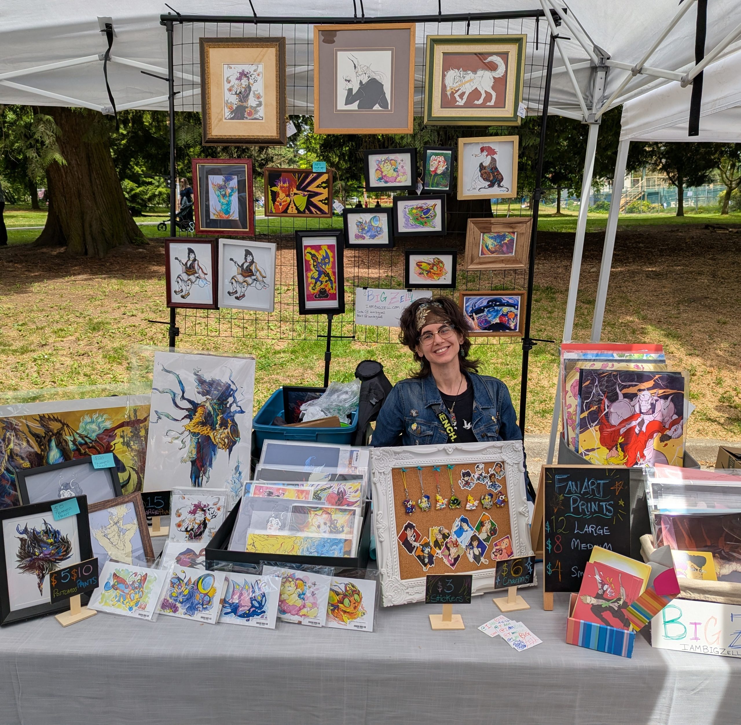 Outdoor artist booth displaying original artwork, framed fantasy illustrations, art prints, stickers, keychains, and a corkboard of colourful character charms.