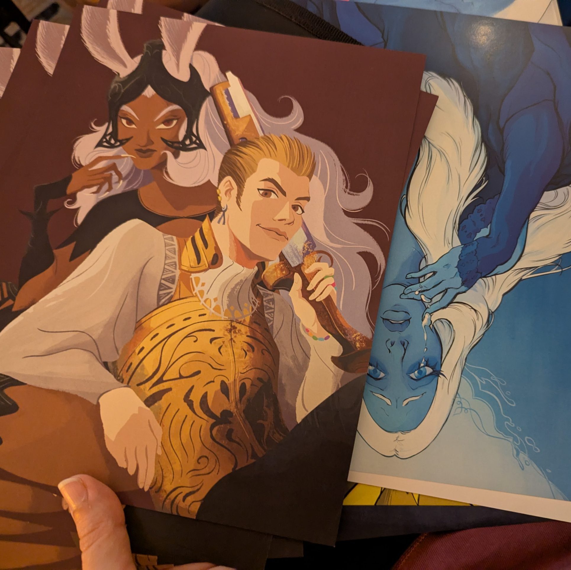 Close-up of several fantasy character art prints, including a white-haired horned woman and a blond man on the left, plus a blue-skinned figure with flowing white hair on the right.