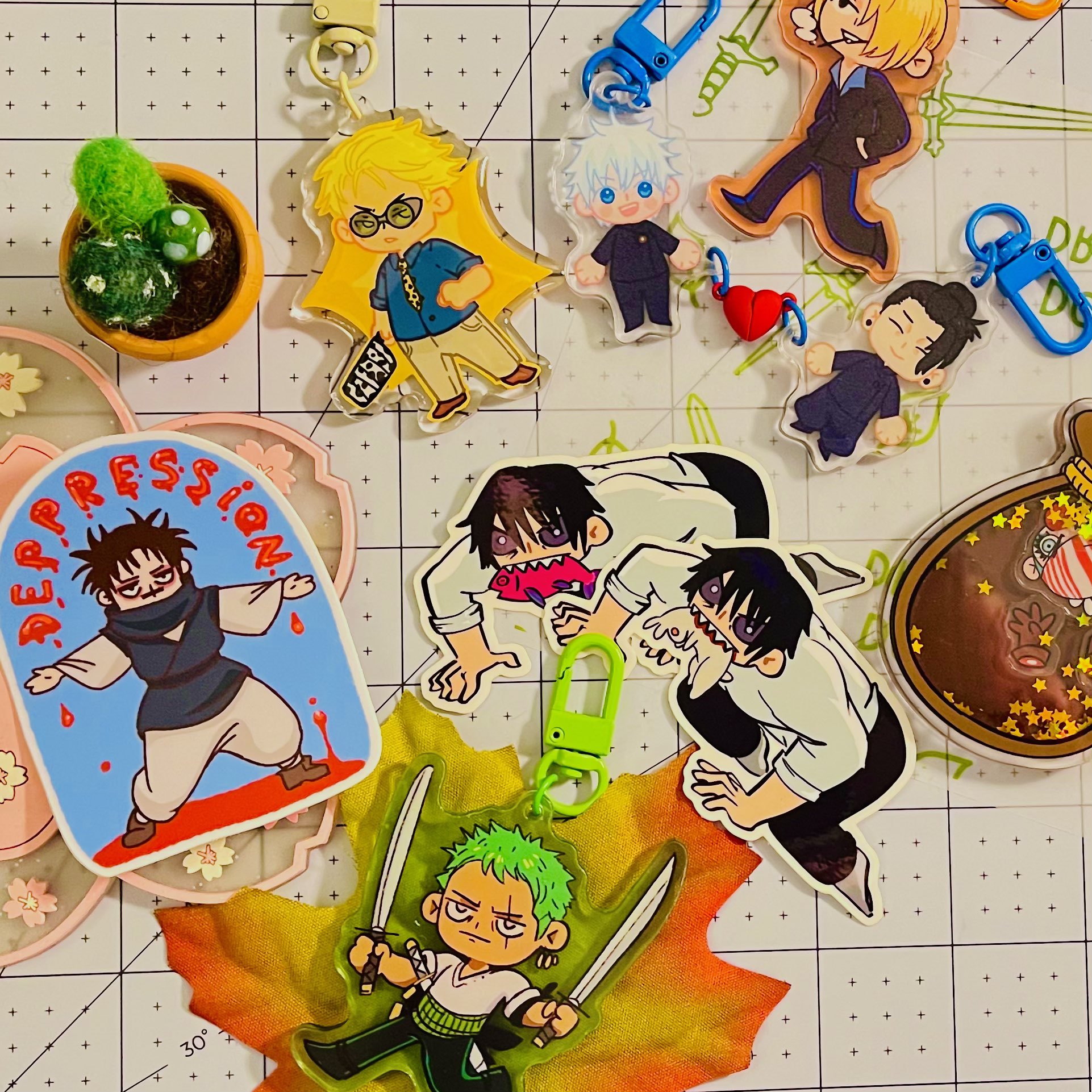 Flat lay of colourful anime-inspired keychains and stickers on a gridded craft mat, including chibi character charms, emotional stickers, washi tape, and a leafy acrylic charm.