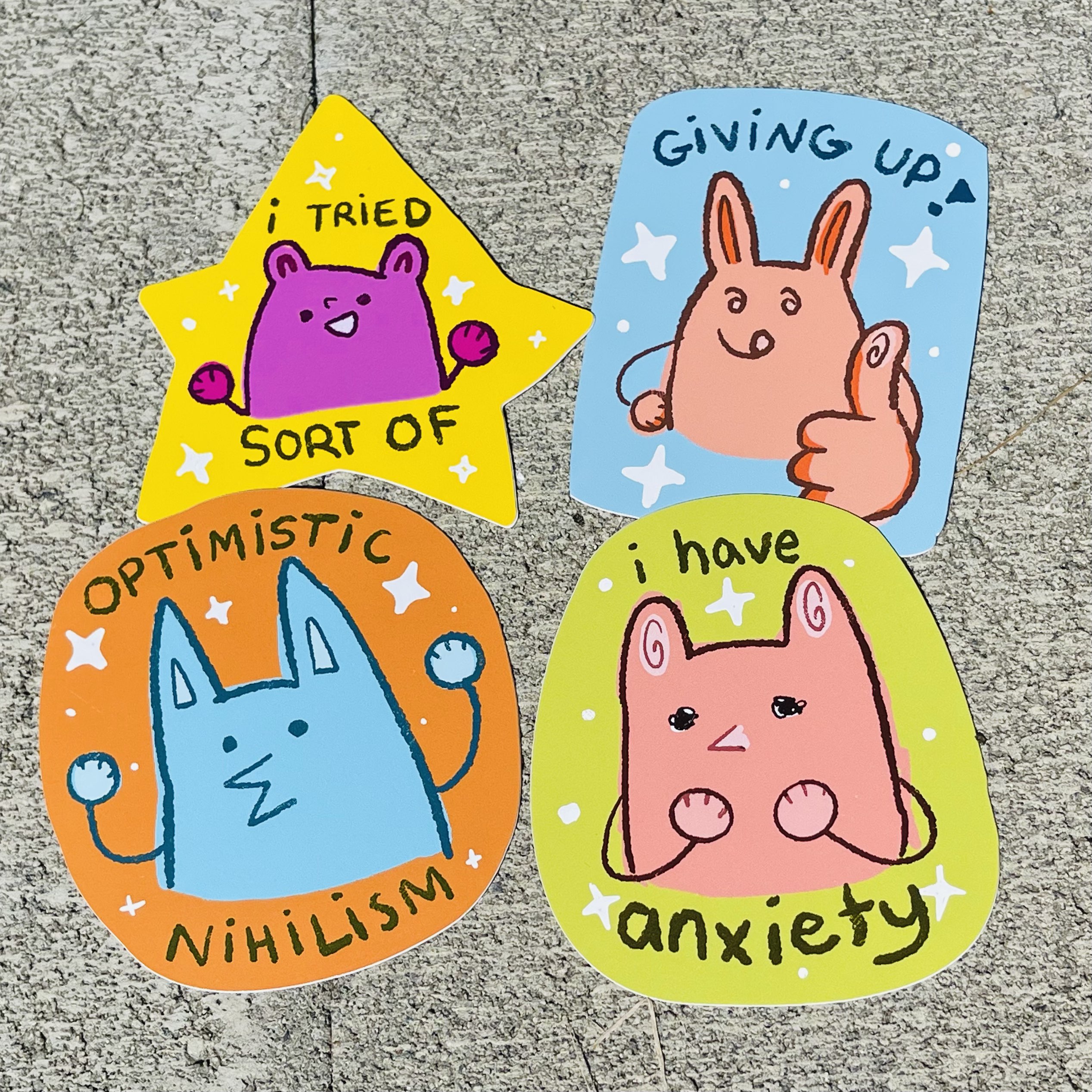 Four bright mental-health themed stickers on concrete reading “I tried sort of,” “giving up,” “optimistic nihilism,” and “I have anxiety,” each with a simple cartoon creature.