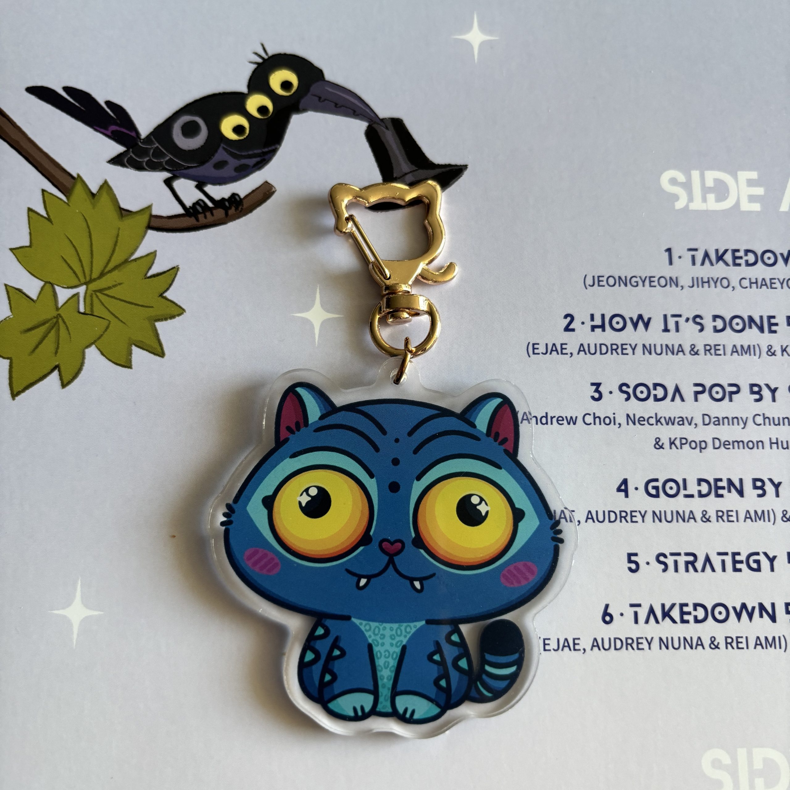Acrylic keychain of a cute blue cat-like character with large yellow eyes, displayed over a printed K-pop themed track list.