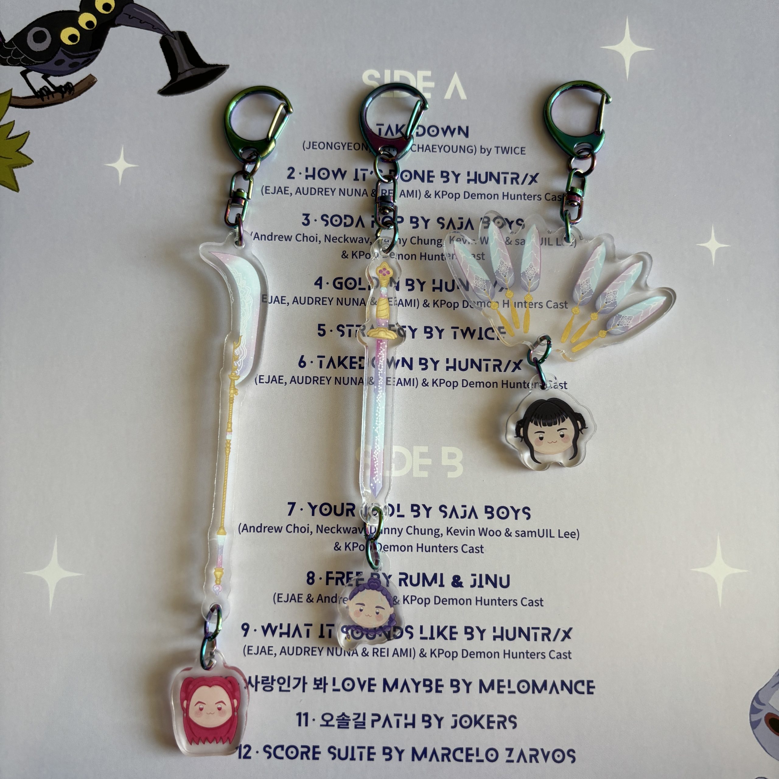 Acrylic K-pop themed keychains with swords, fans, and small character charms arranged on a printed track list background.