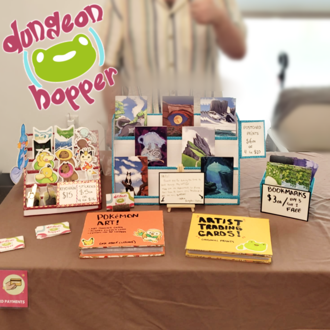 Dungeon Hopper vendor table with stickers, keychains, postcard prints, bookmarks, Pokémon art cards, and artist trading cards.