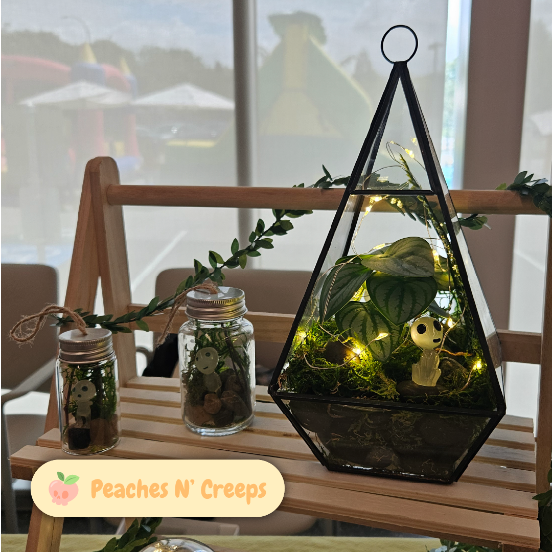 Handmade terrariums by Peaches N' Creeps with small forest spirit figures, moss, plants, stones, and warm fairy lights displayed on a wooden shelf.