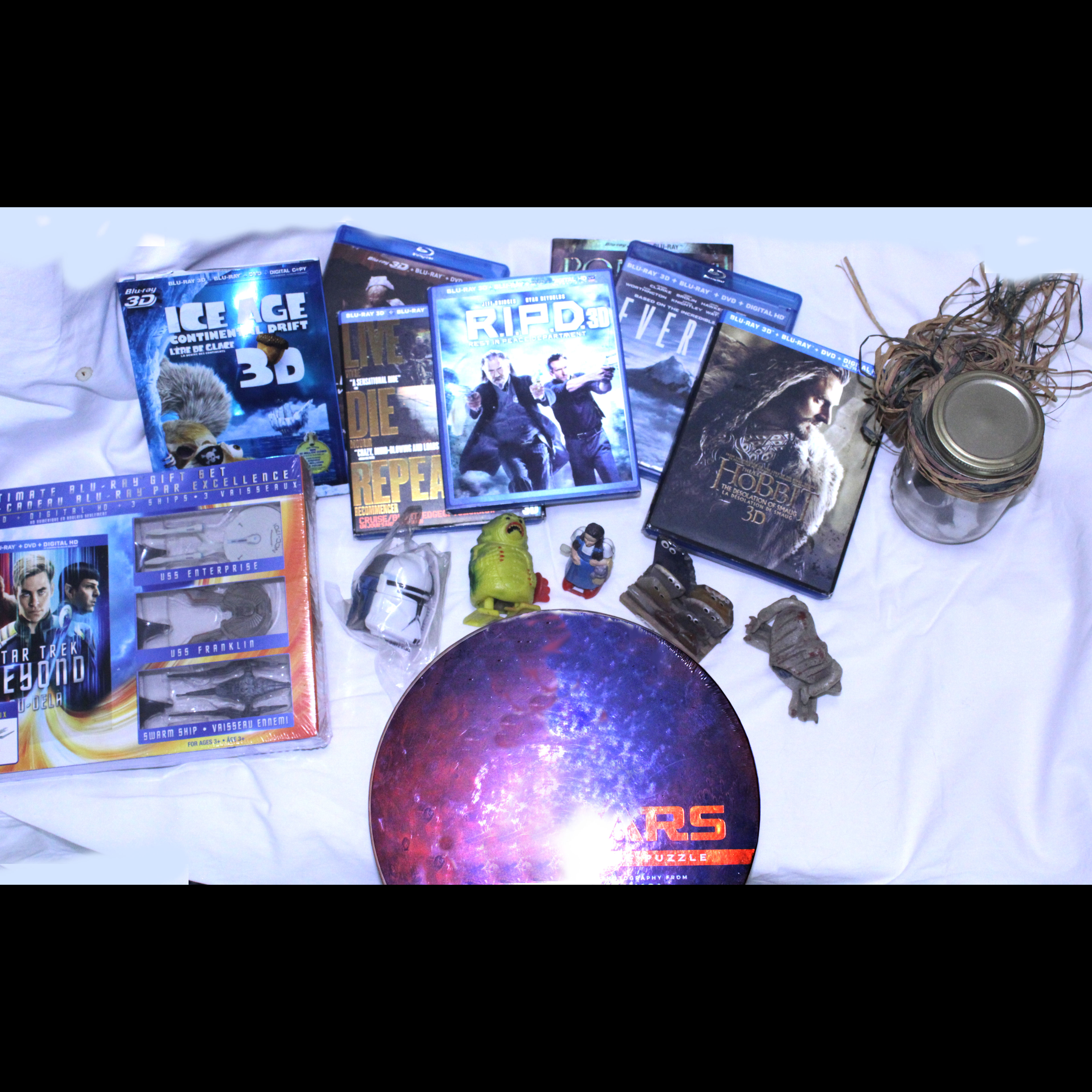 Assortment of sci-fi and fantasy collectibles on a white cloth, including 3D Blu-rays, Star Trek mini ships, small figures, a Mars puzzle, and a glass jar prop.