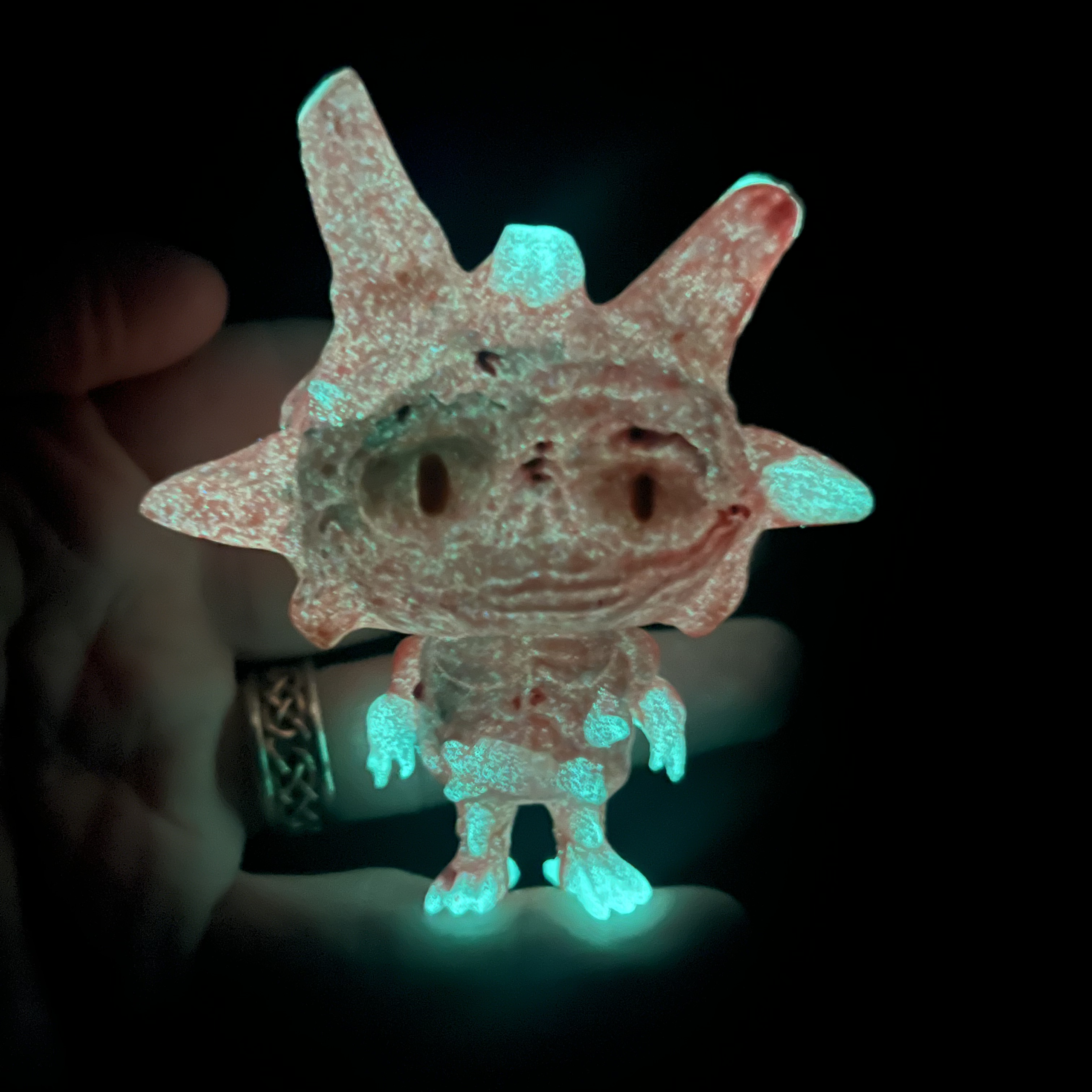 Glow-in-the-dark hand-cast monster toy with small horns, big eyes, and turquoise glowing hands and feet, held against a dark background.