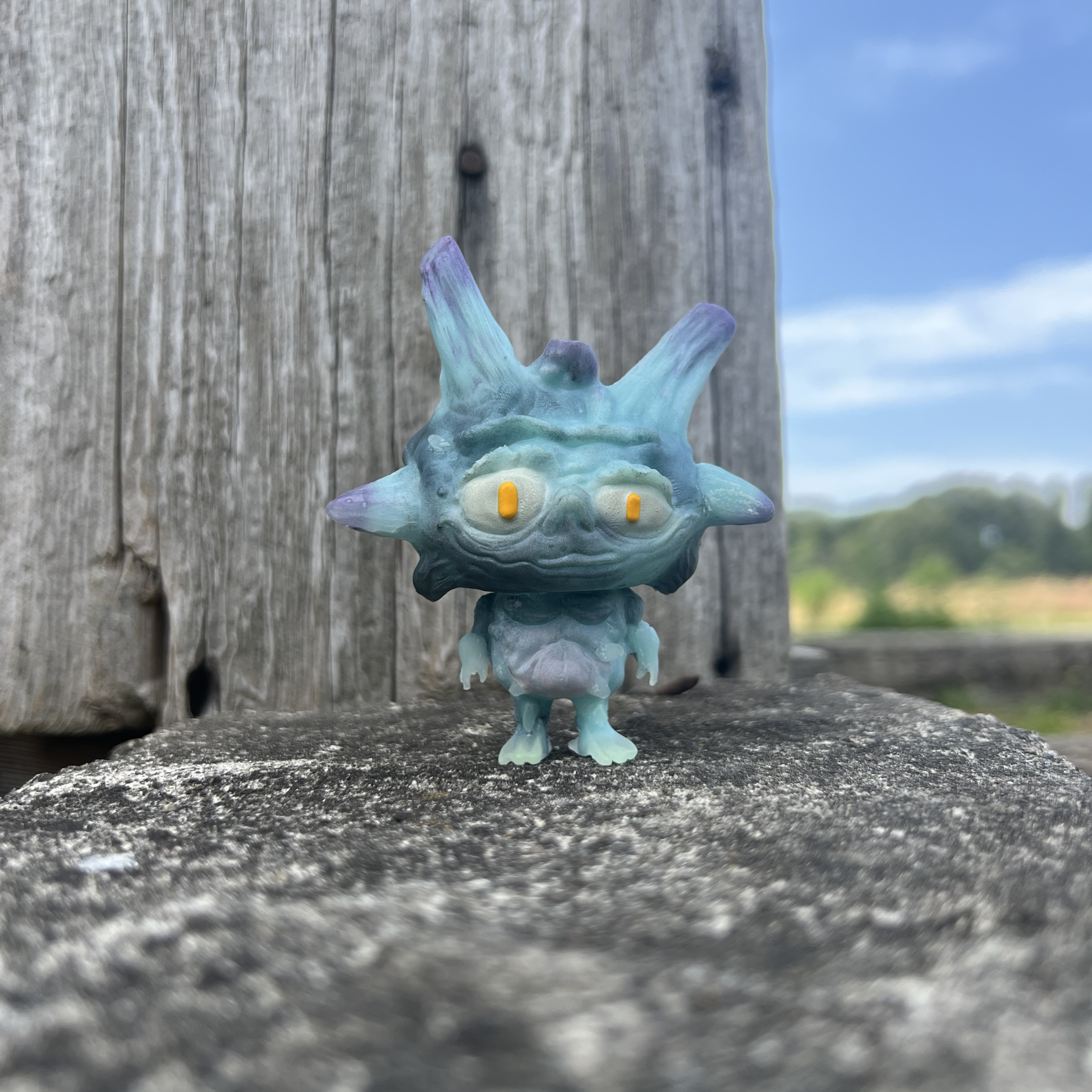 Hand-painted blue monster toy with yellow eyes and purple-tipped horns posed outdoors on a wooden surface.