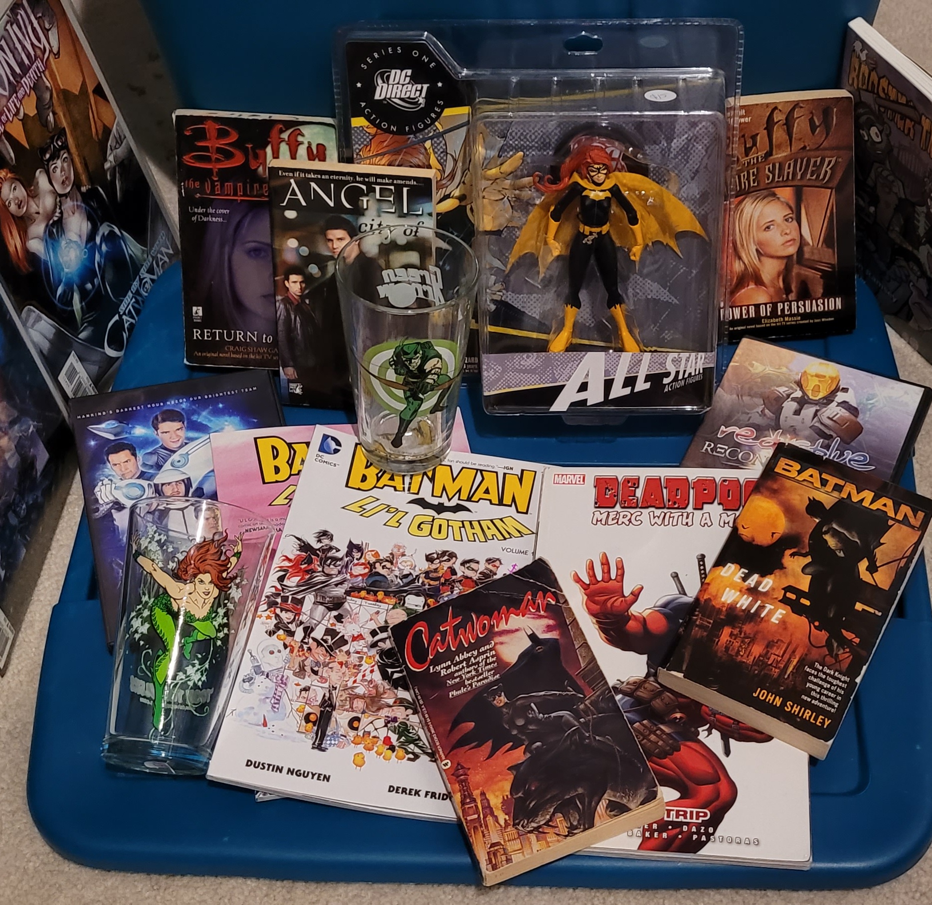 Assortment of geeky collectibles on a blue surface, including Buffy and Angel books, Batman graphic novels, a Deadpool book, a Batgirl action figure, and superhero-themed glasses.