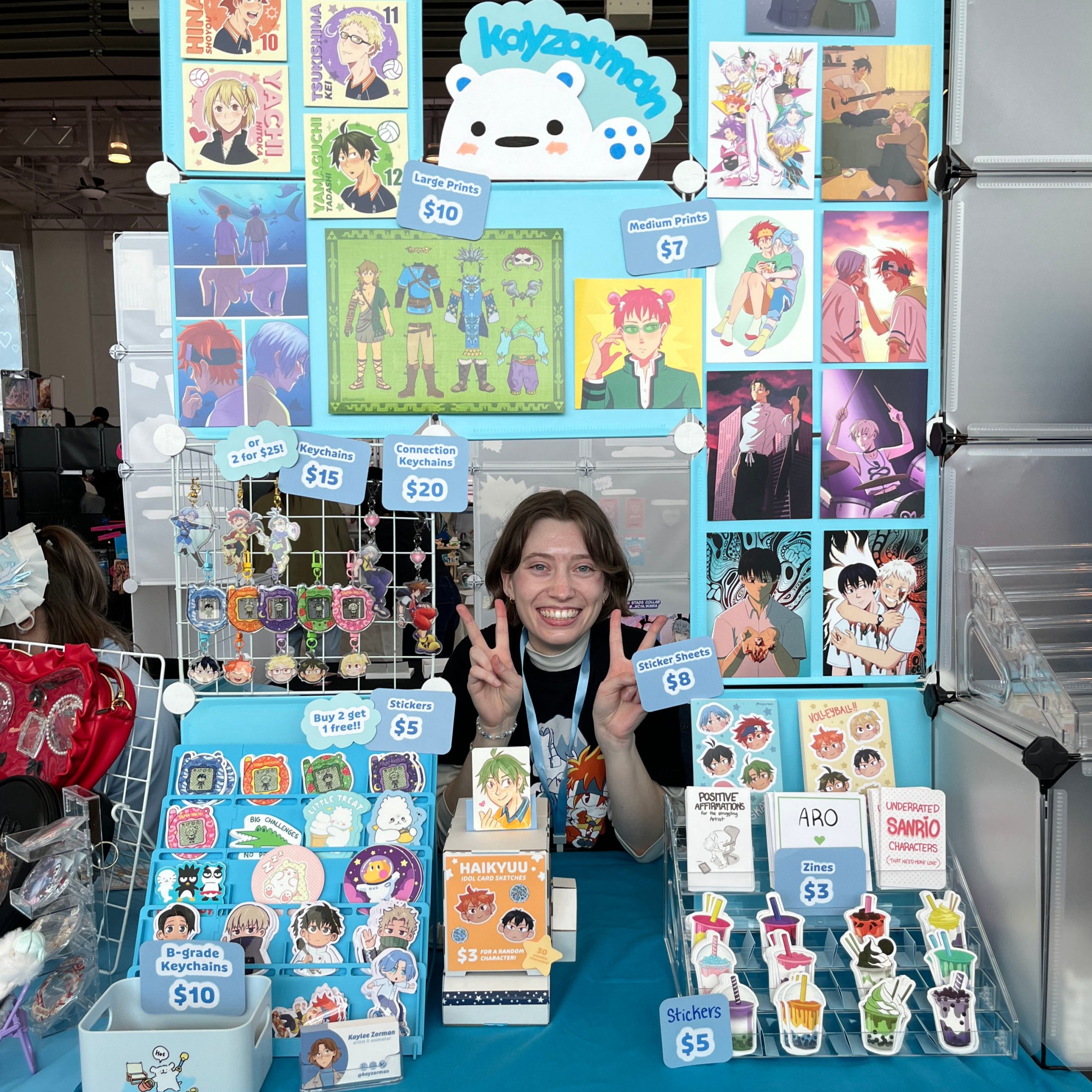 Anime artist table display with colourful prints, keychains, stickers, sticker sheets, zines, and price signs, with the vendor smiling behind the booth.