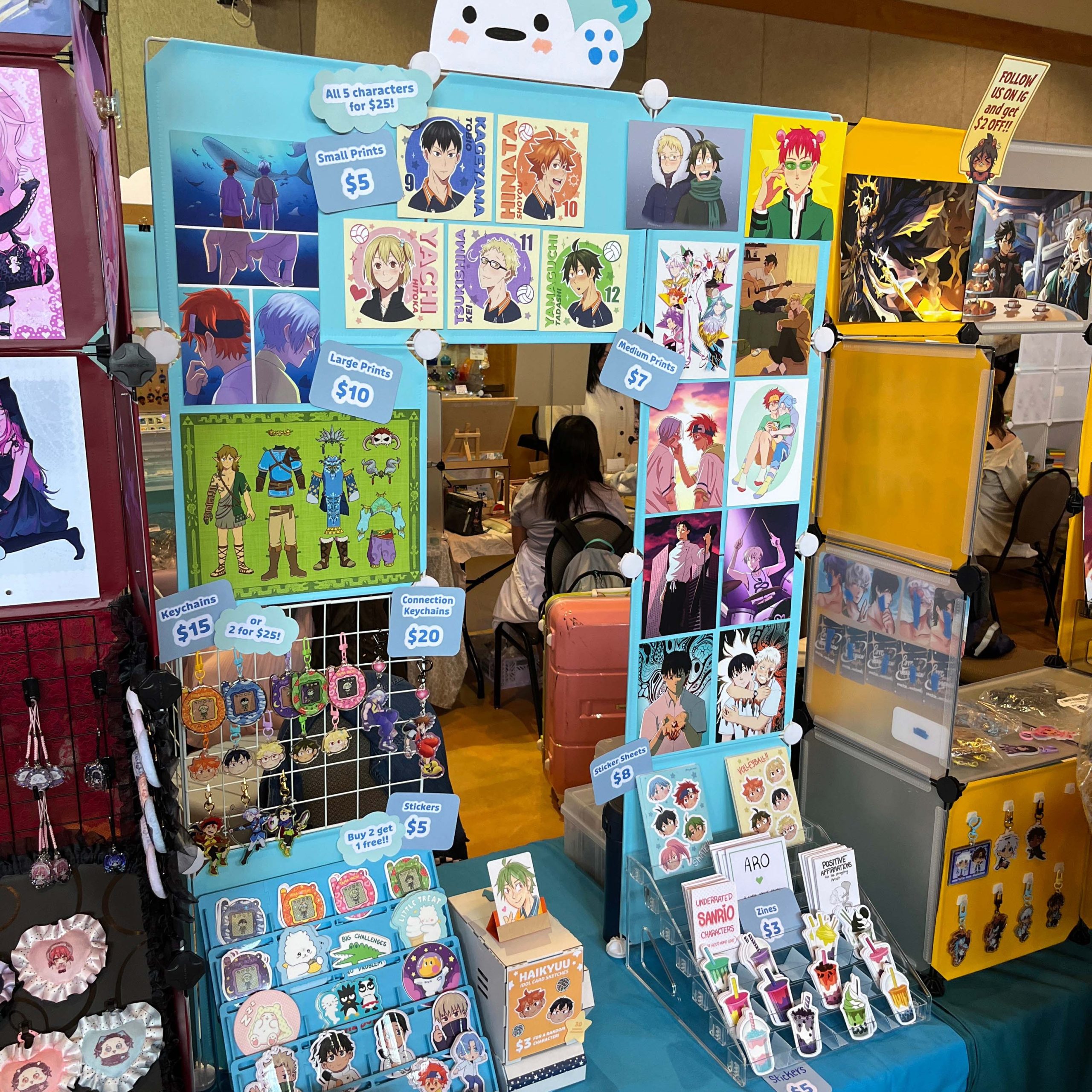 Bright blue and yellow anime merchandise booth displaying prints, keychains, stickers, zines, and sticker sheets from fandoms including Haikyuu and SK8.