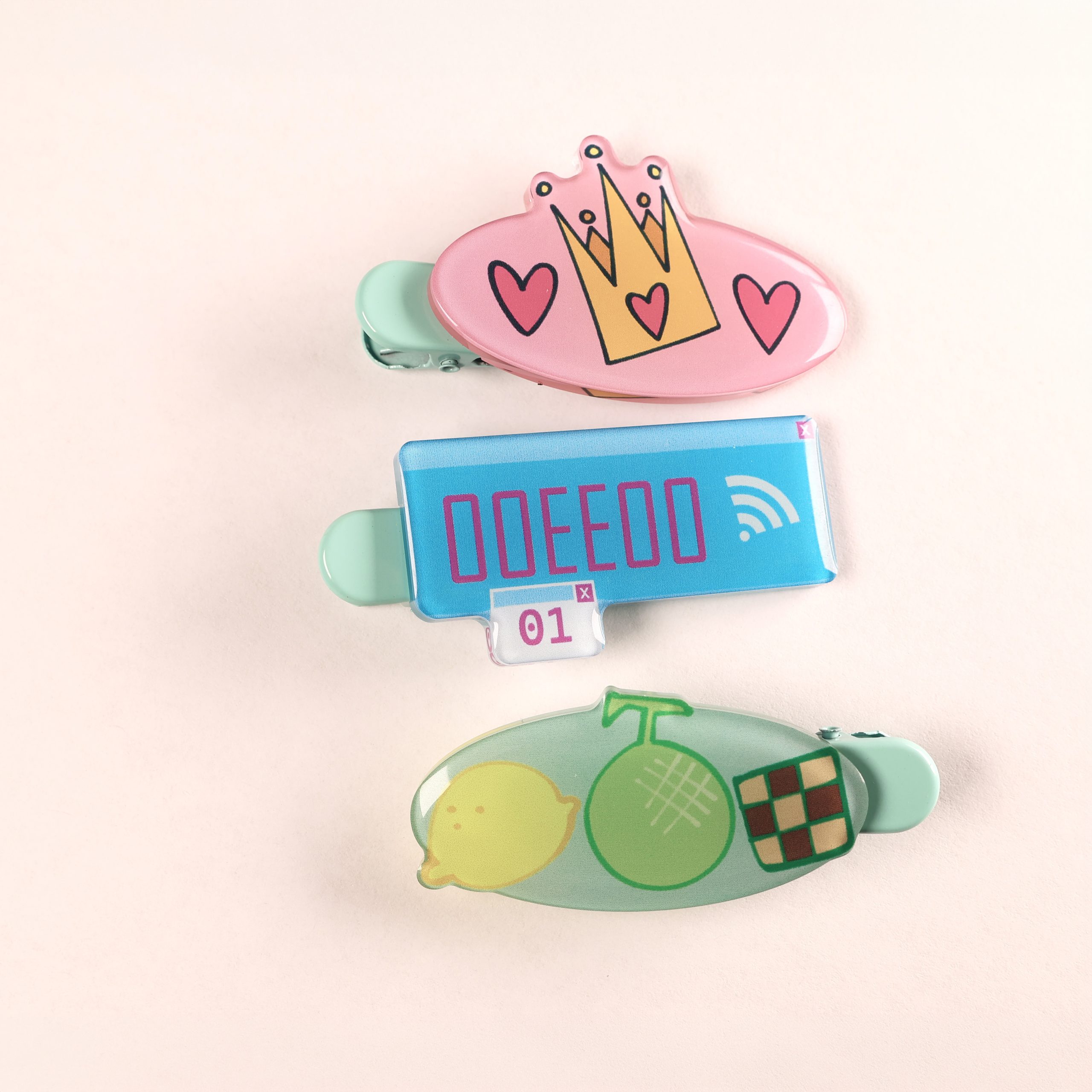 Three handmade hair clips on a pale background, including a pink crown design, a blue digital-style design, and a green clip with lemon, melon, and chocolate motifs.