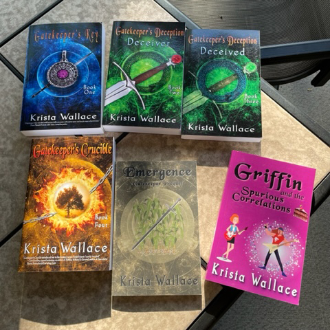 Six Krista Wallace fantasy books arranged on a tabletop, including The Gatekeeper series, the Emergence prequel novella, and the pink-covered Griffin and the Spurious Correlations.