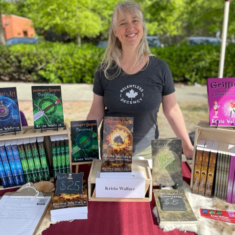 Fantasy author Krista Wallace smiling behind an outdoor vendor table displaying her books, with copies of The Gatekeeper series, Emergence, and Griffin and the Spurious Correlations.