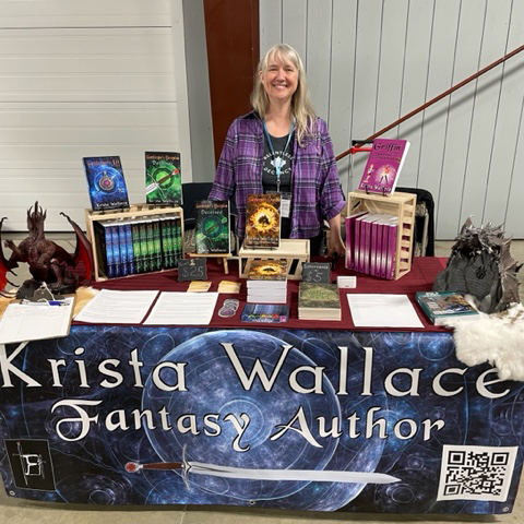 Krista Wallace standing behind a convention table with fantasy books, display stands, dragon decorations, and a large “Krista Wallace Fantasy Author” banner.