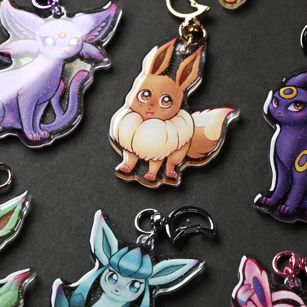 Close-up of glittery acrylic keychains featuring cute Pokémon-inspired characters, including Eevee, Espeon, Umbreon, Glaceon, and other designs.