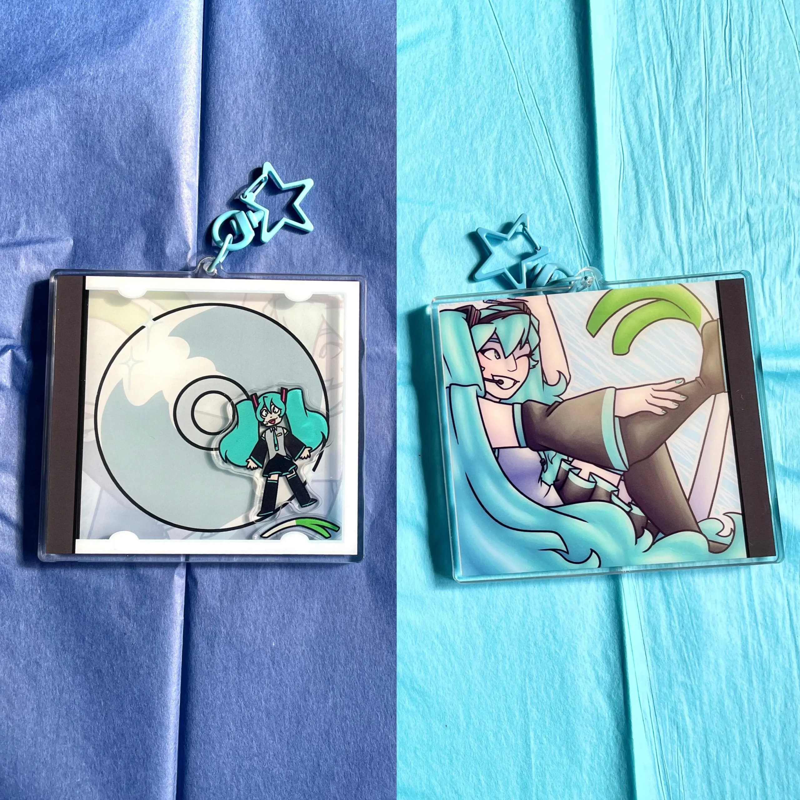 Two acrylic keychains on blue tissue paper featuring a teal-haired anime-style character, one with a CD design and one with a close-up character illustration.