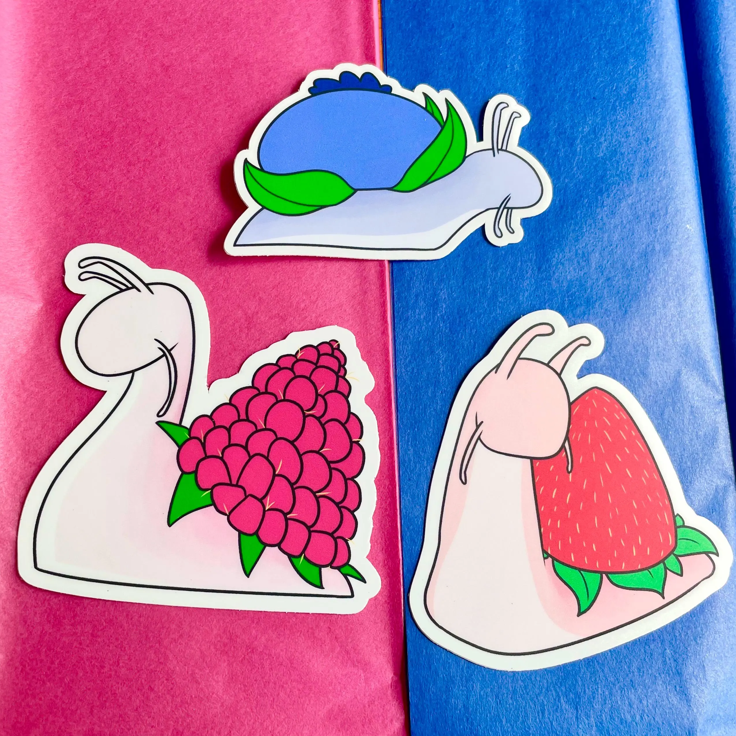 Three cute snail stickers on pink and blue tissue paper, including snails with a hydrangea, blueberry, and strawberry shell.