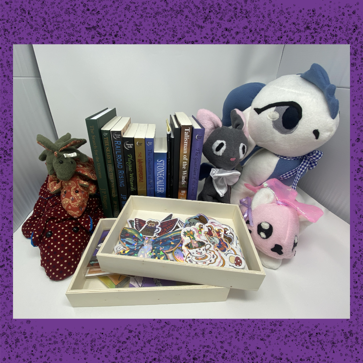 Display of fantasy books, plush animals, and sticker artwork arranged together, including dragon plushies, cat plushies, and a tray of colourful stickers.