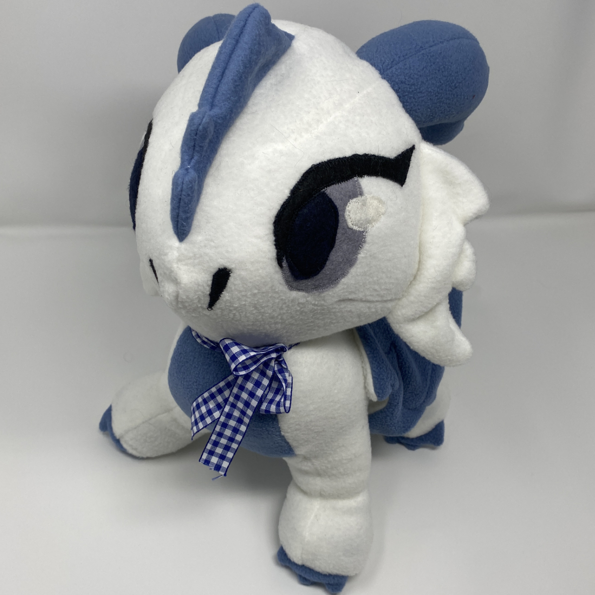 Blue and white handmade plush creature with large embroidered eyes and a checkered bow, posed against a plain grey background.