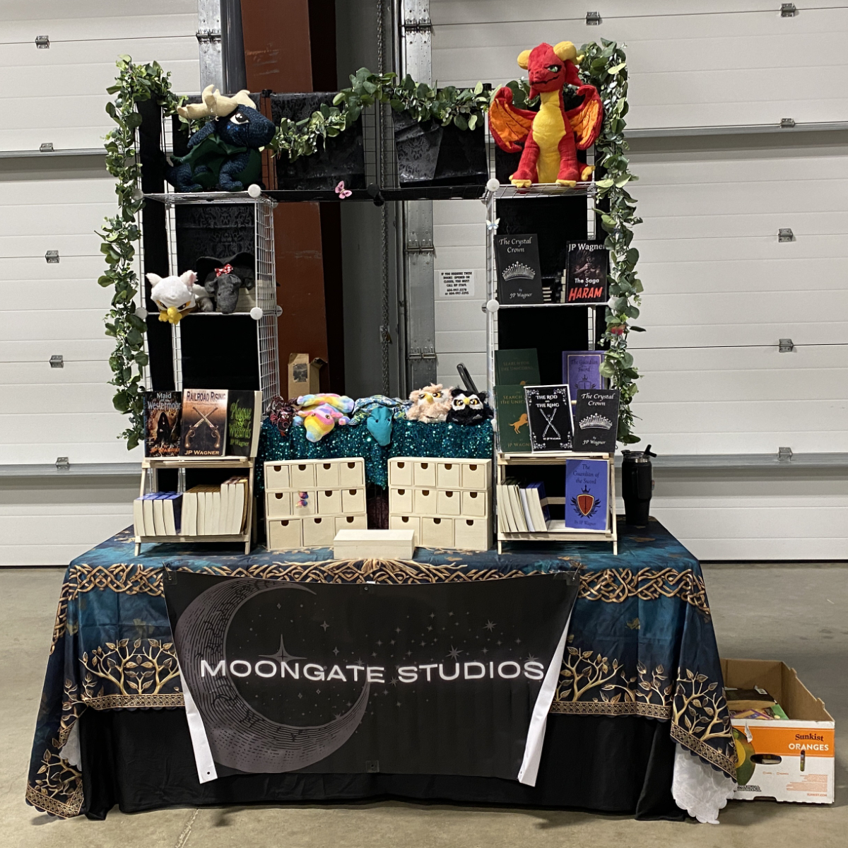 Moongate Studios vendor table displaying books, plush dragons and animals, small drawers, greenery, and a large Moongate Studios banner.