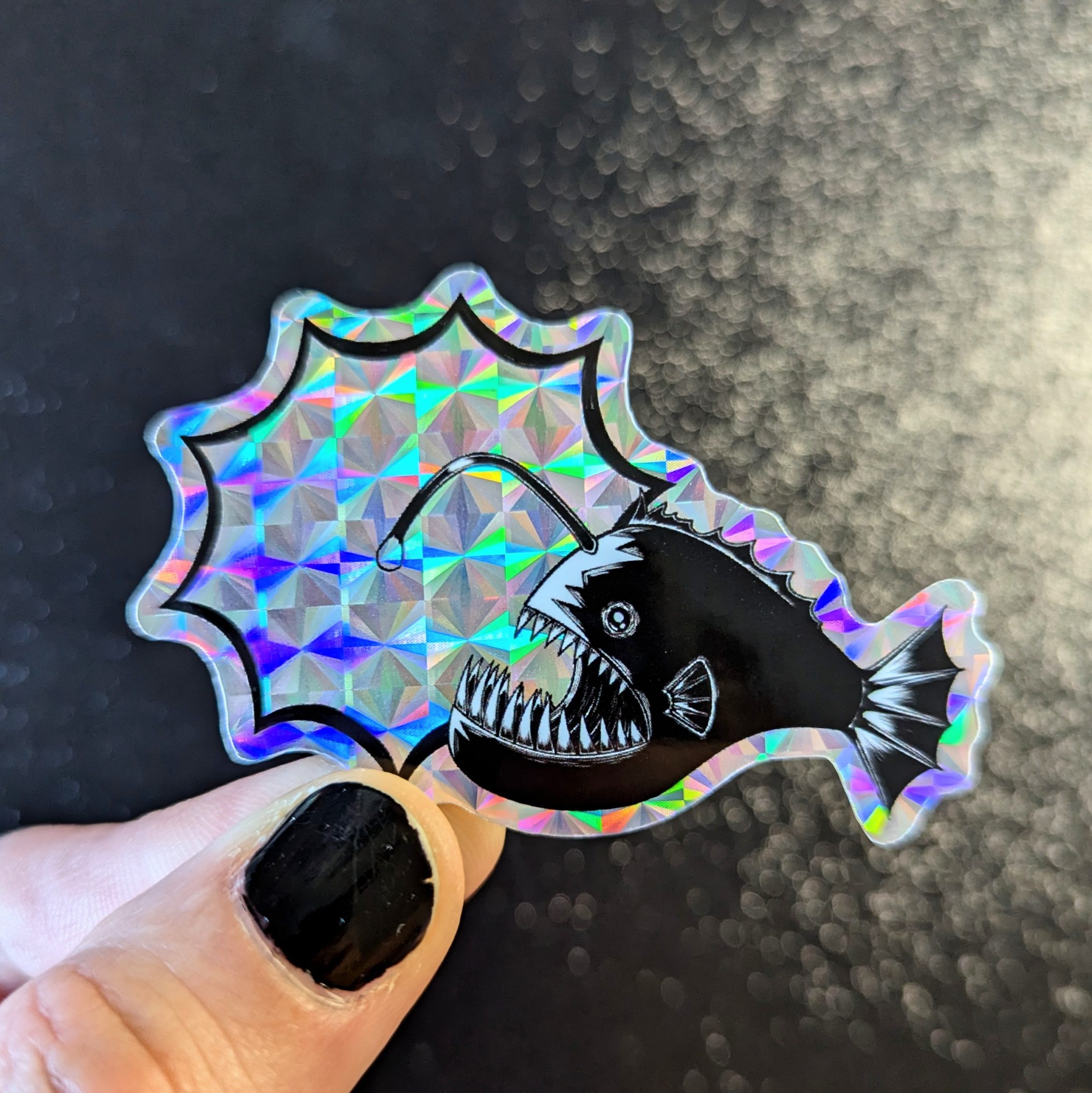Holographic sticker of a black anglerfish with sharp teeth and a glowing lure, held against a dark textured background.