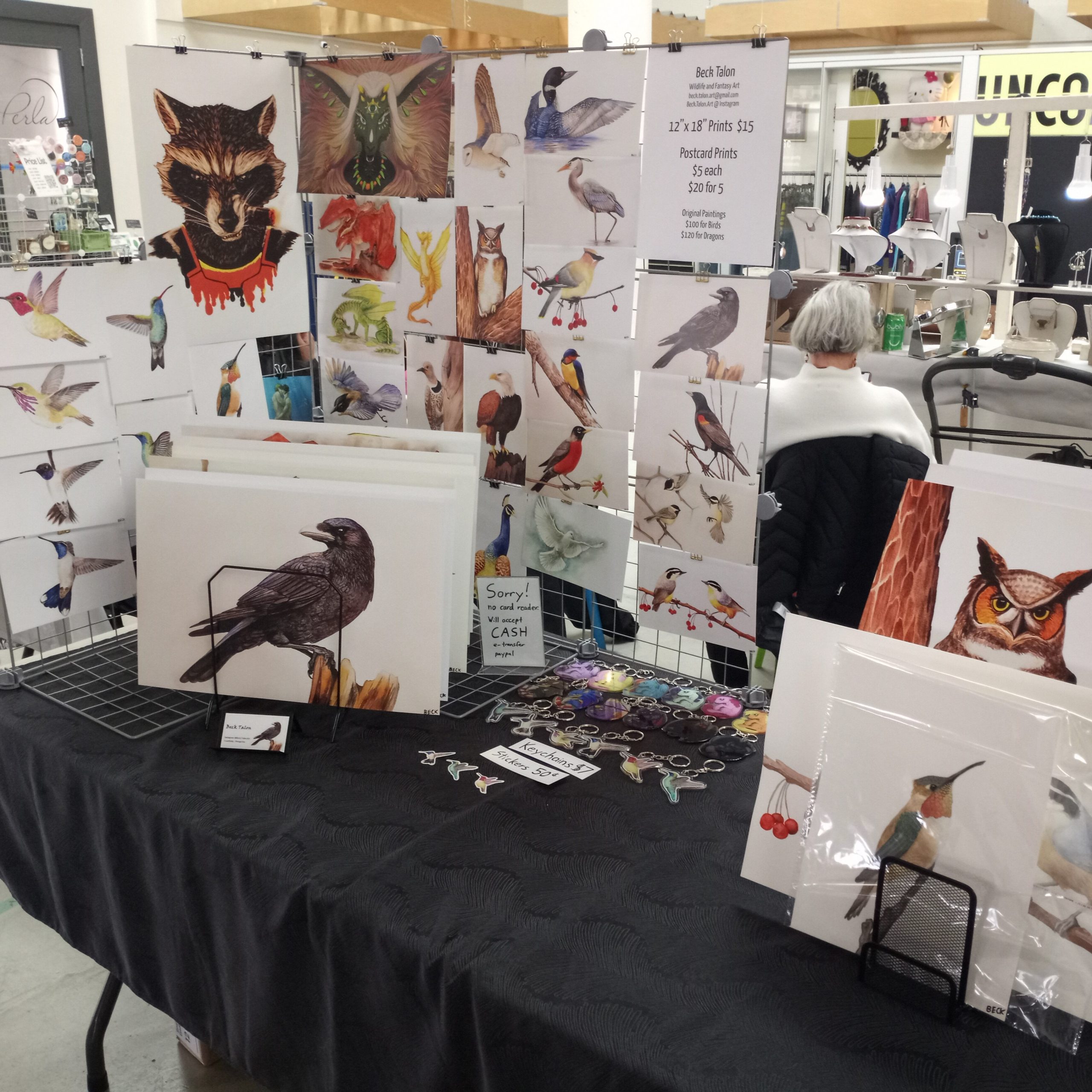 Artist table display featuring watercolour and digital wildlife and fantasy art prints, including birds, a raven, owl, raccoon, dragons, keychains, and stickers.