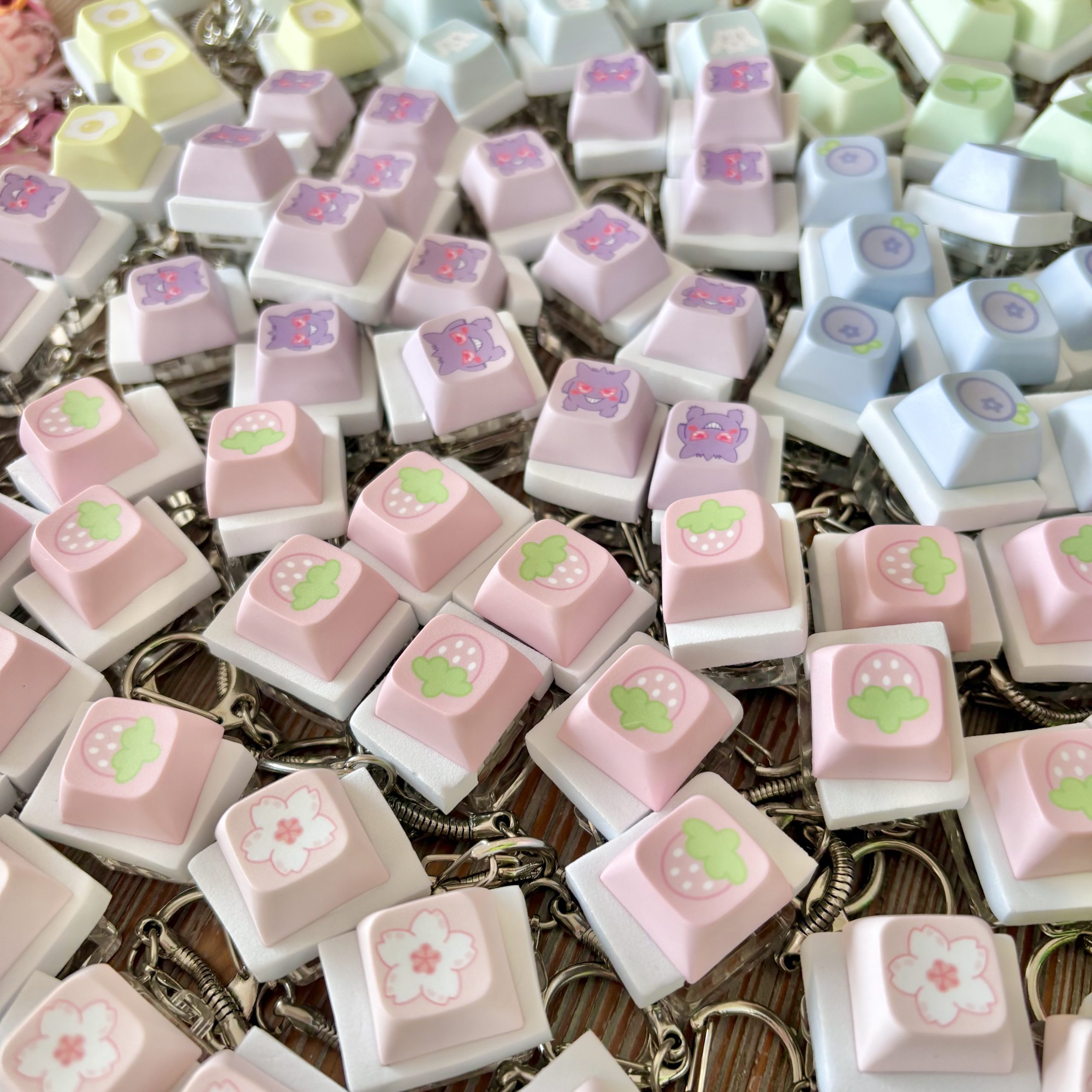 Close-up of pastel keyboard keycap keychains with cute designs including strawberries, cherry blossoms, ghost faces, blueberries, eggs, and green leaves.