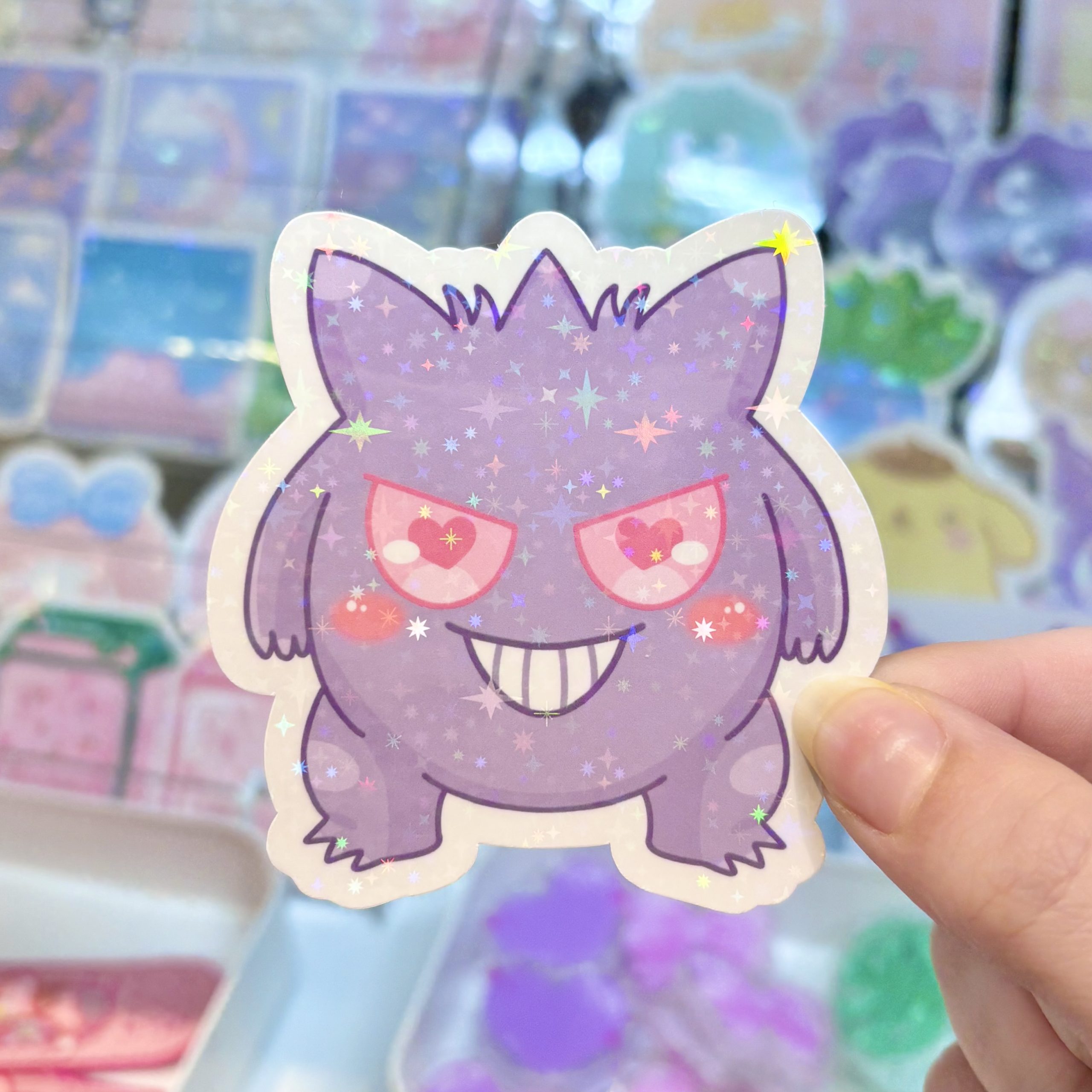 Glittery sticker of a cute purple ghost-like character with pink eyes and a big grin, held in front of a pastel merchandise display