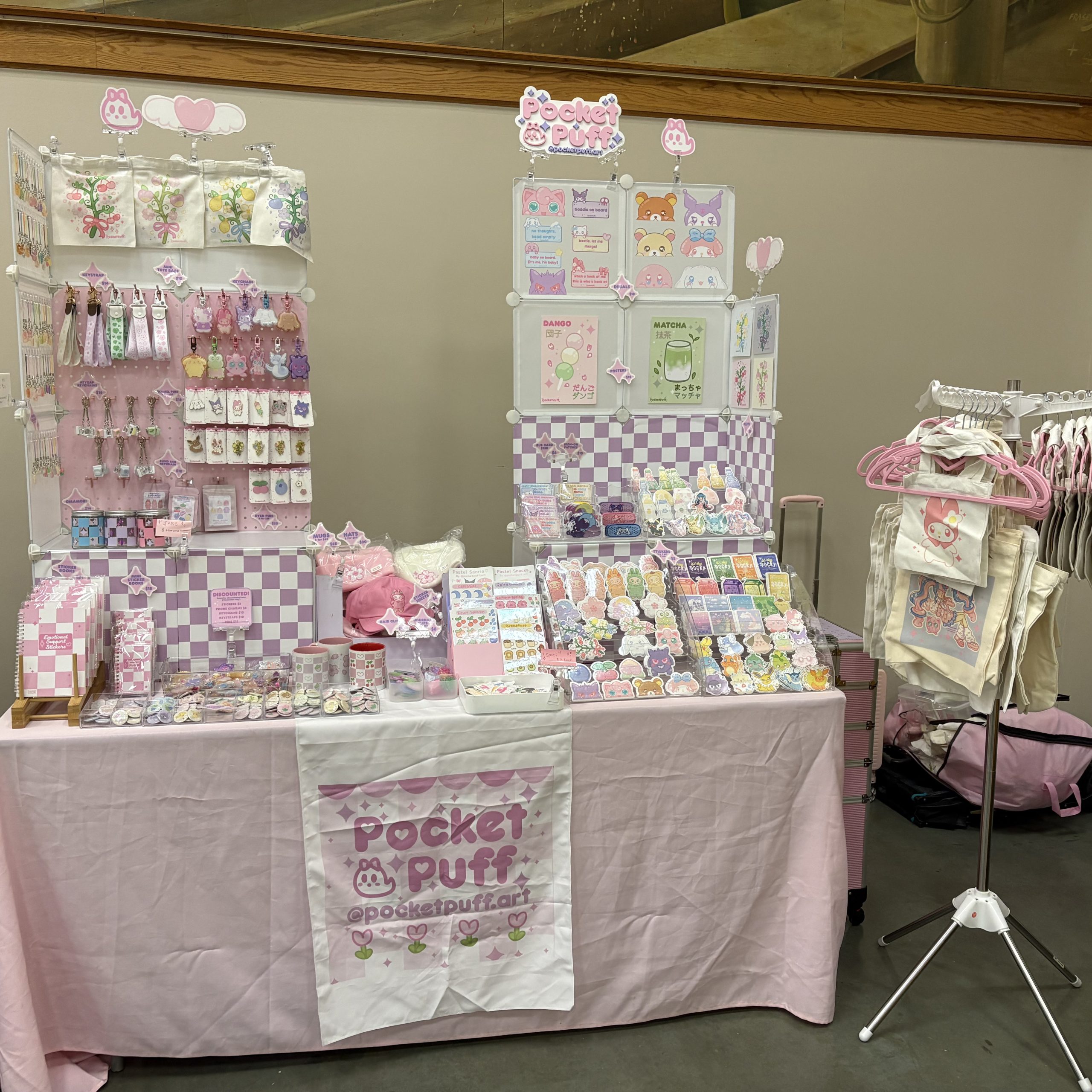 Pastel PocketPuff vendor table with stickers, keychains, charms, pins, prints, bags, and accessories in a pink-and-purple kawaii displa