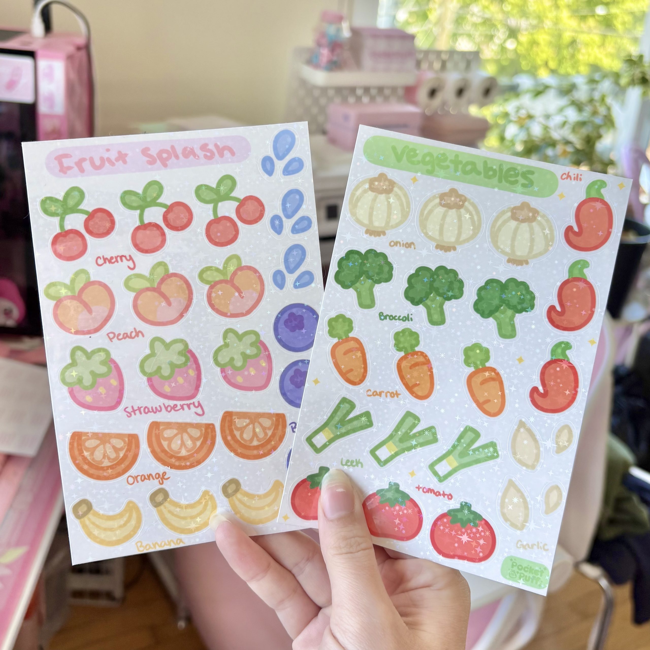 Two sparkly sticker sheets held in front of a bright window, featuring cute fruit and vegetable designs such as cherries, strawberries, bananas, onions, broccoli, carrots, leeks, and toma