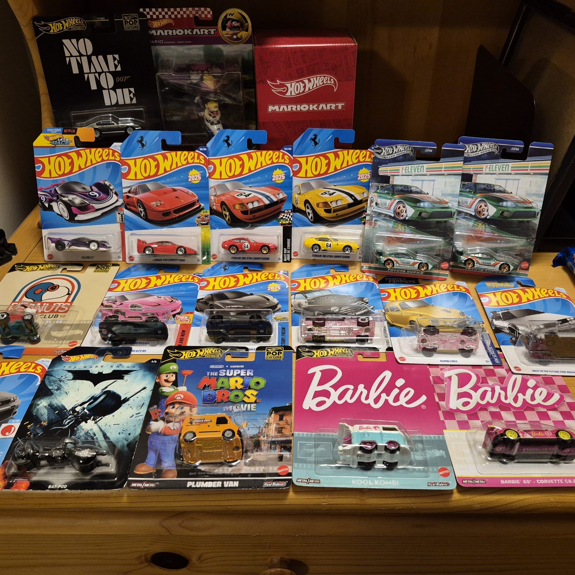 Assorted packaged Hot Wheels toy cars displayed on a wooden desk, including themed cars from Mario Kart, Barbie, Batman, Back to the Future, and other collector series.