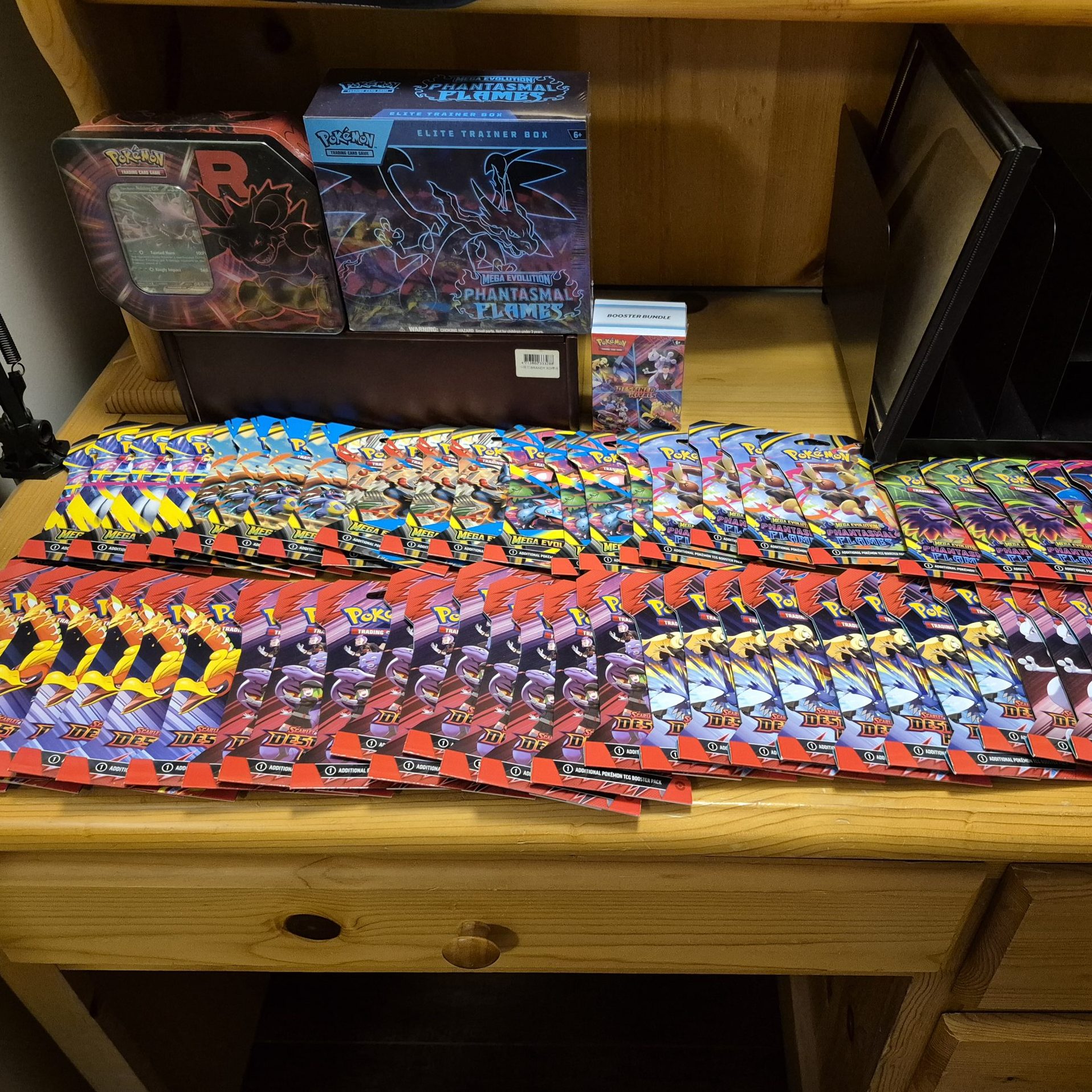 Large display of sealed Pokémon trading card booster packs spread across a wooden desk, with Pokémon tins, booster bundles, and an Elite Trainer Box behind them.