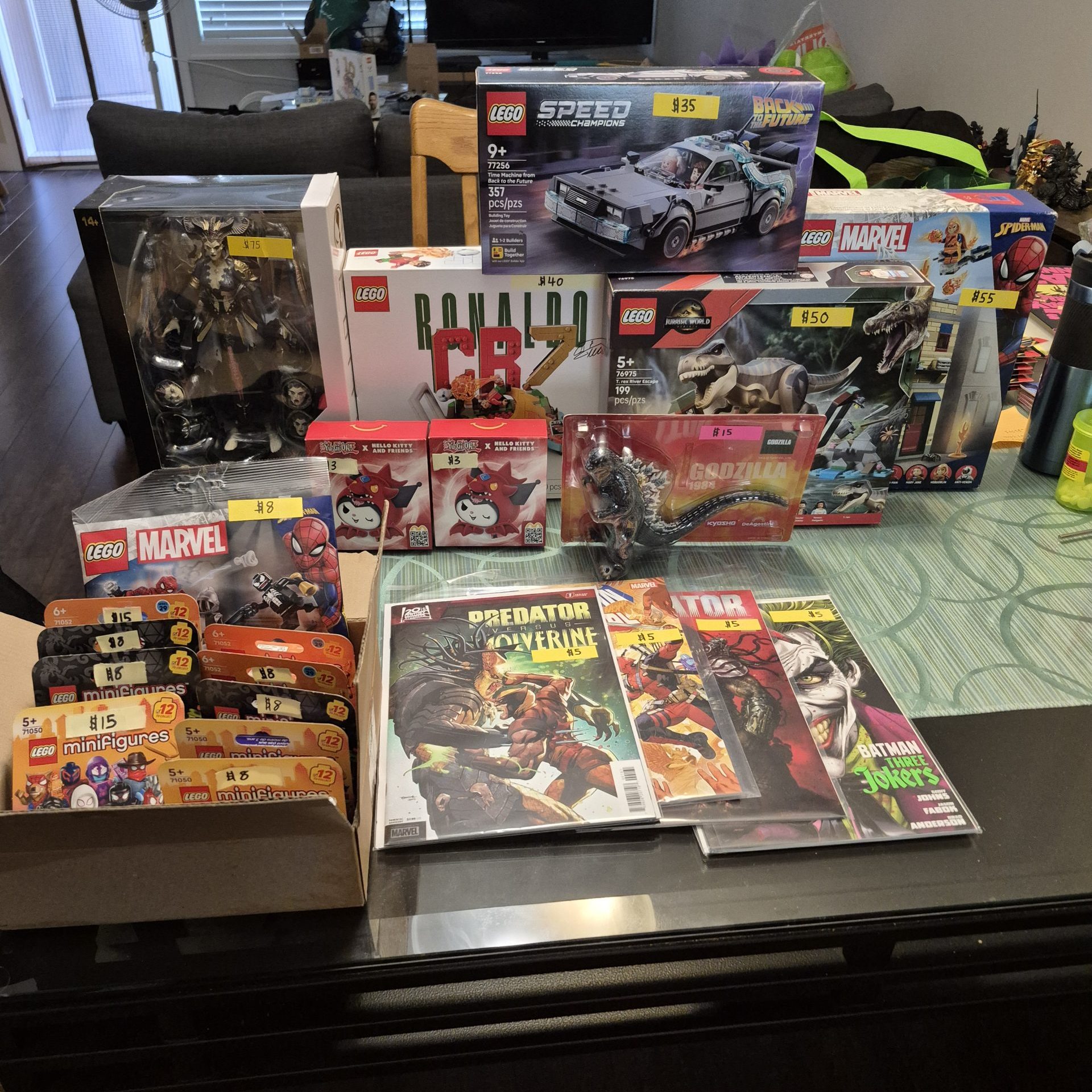 Table display of collectibles for sale, including LEGO sets and minifigure packs, boxed figures, Godzilla merchandise, and comic books featuring Predator, Wolverine, Batman, and other characters