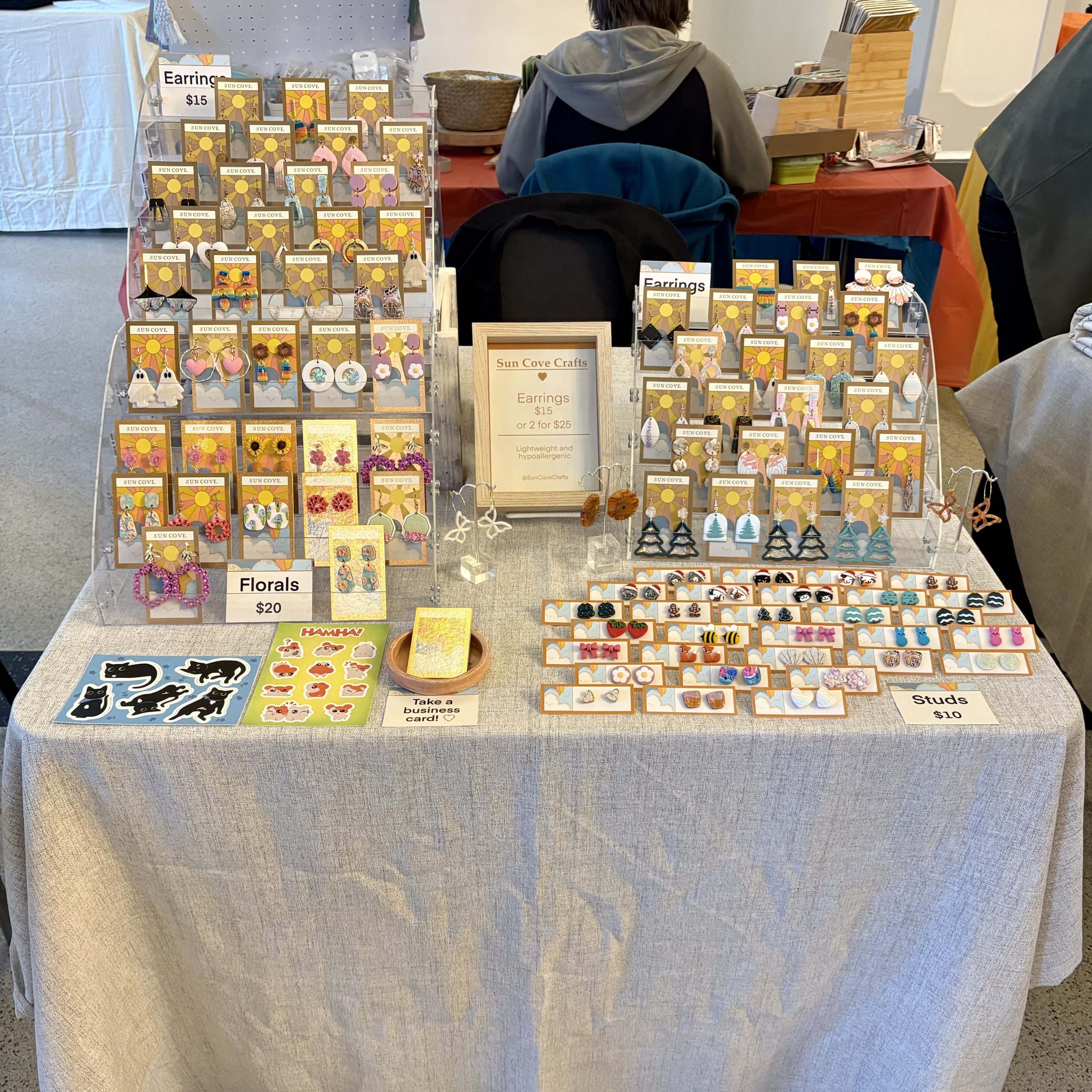 Sun Cove Crafts vendor table display with handmade polymer clay earrings, floral designs, studs, stickers, and small signage on a neutral tablecloth.