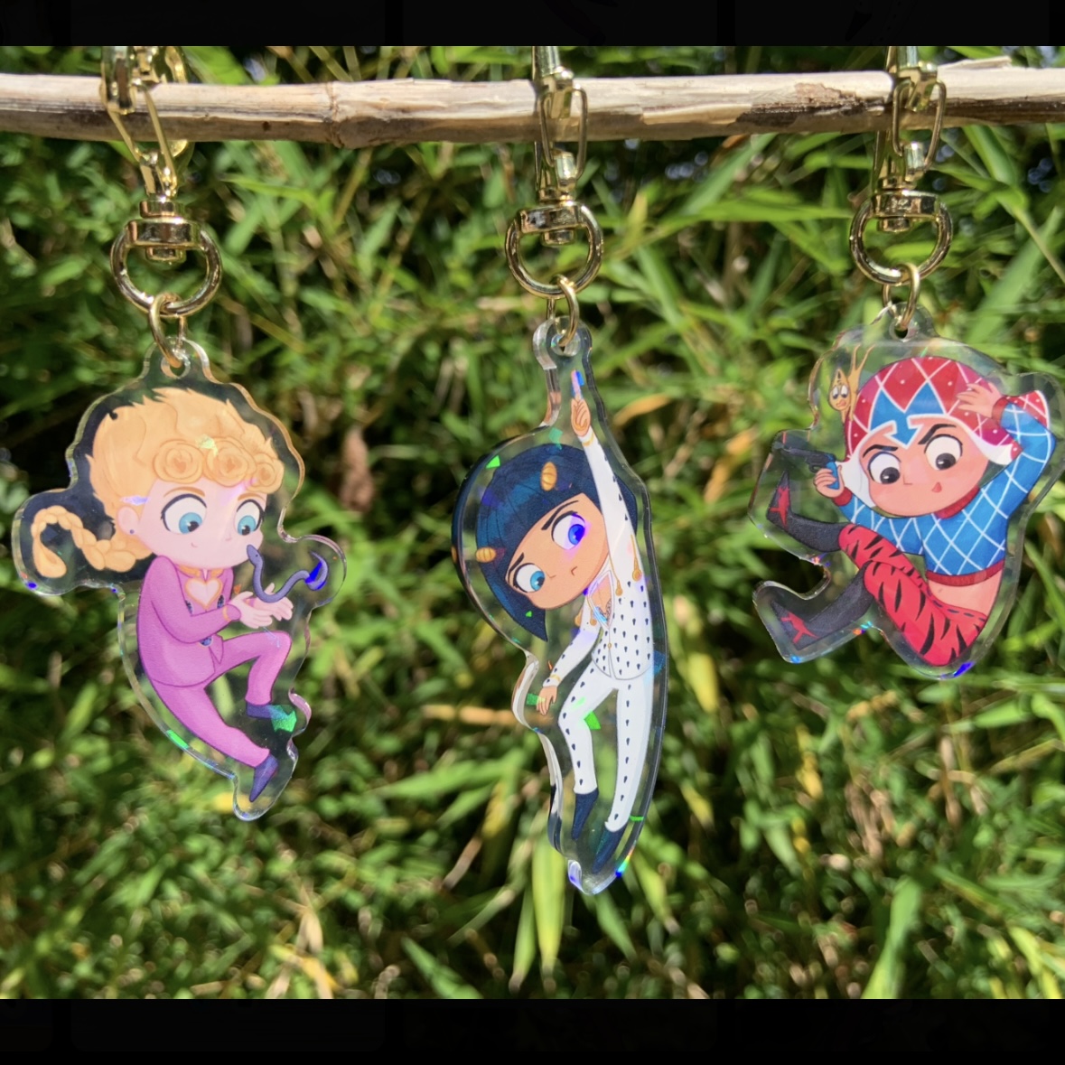 Three colourful acrylic fandom charms hanging from a branch outdoors, featuring cute character art with gold hardware.