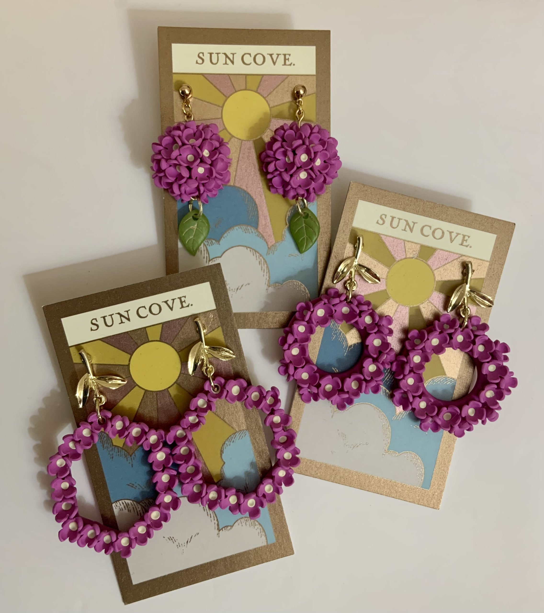 Three pairs of Sun Cove Crafts floral polymer clay earrings displayed on branded backing cards with sun and cloud artwork.