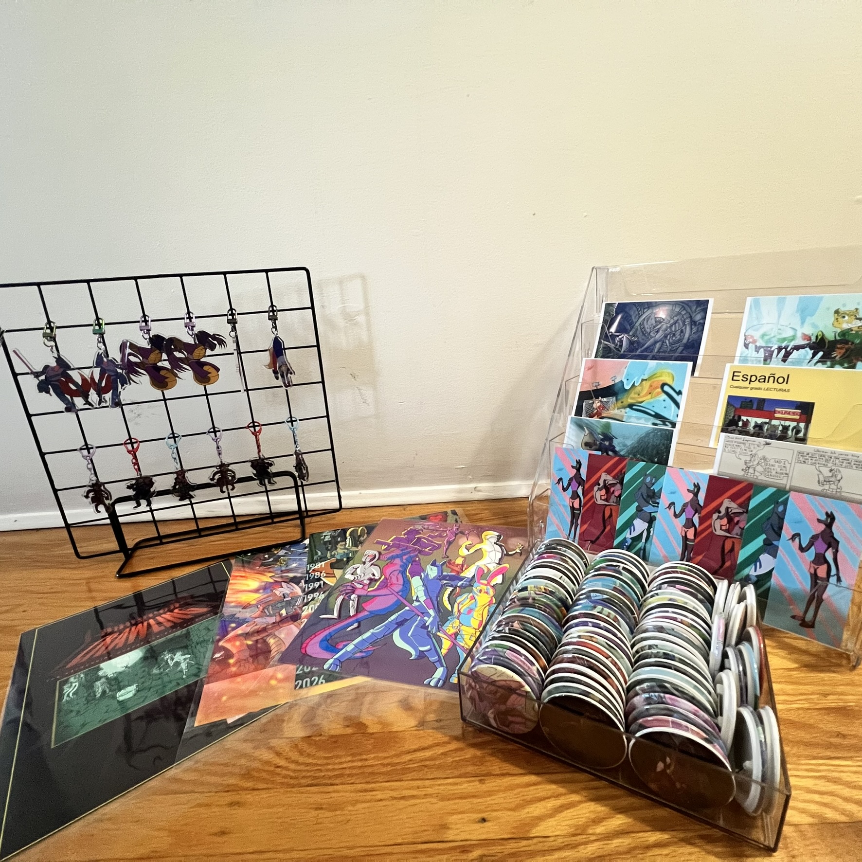 Table display with acrylic charms, art prints, rows of buttons, postcards, and original comics arranged on a wooden surface.